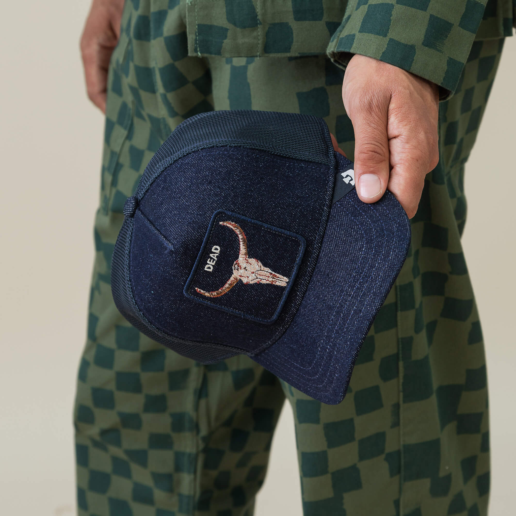 A person in a green checkered outfit holds the Goorin Bros. Denim Dead cap—a navy blue biker hat featuring a bull skull embroidery patch and the word "DEAD.