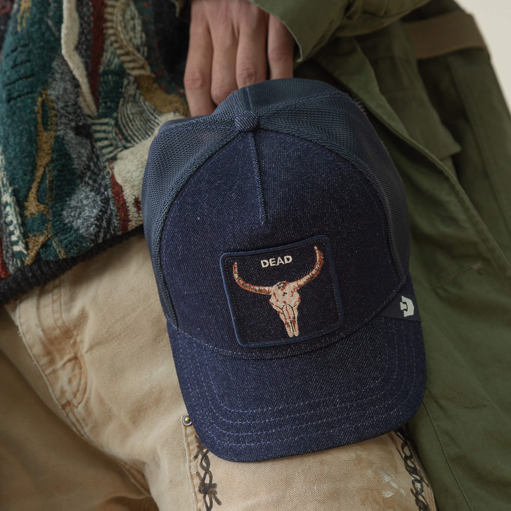 A person holds the Goorin Bros. Denim Dead trucker hat, featuring a bold bull skull patch with "DEAD" embroidered above it.