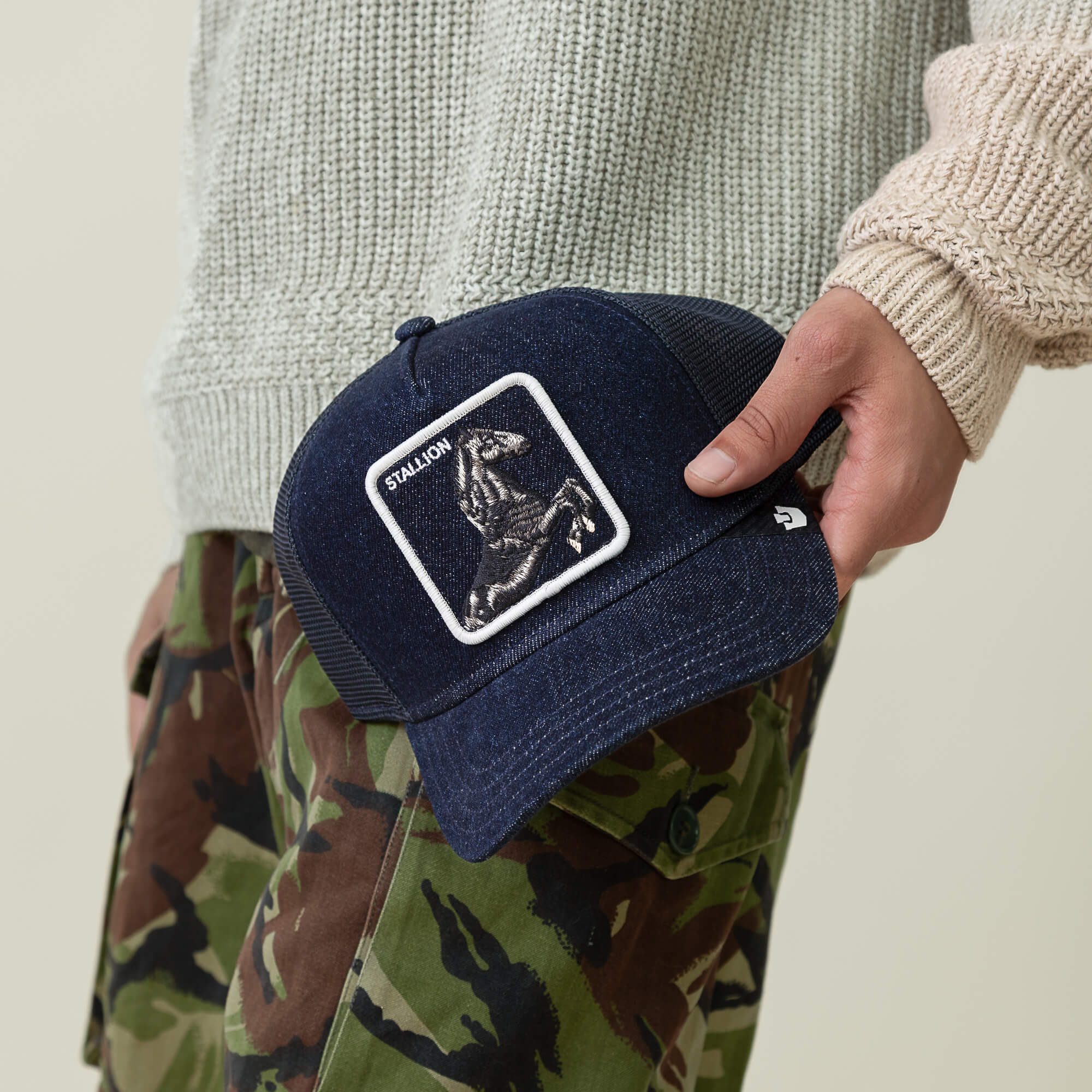 A person holds the Goorin Bros. Denim Stallion cap, featuring an embroidered horse patch, paired with a knit sweater and camouflage pants for a laid-back arena look.