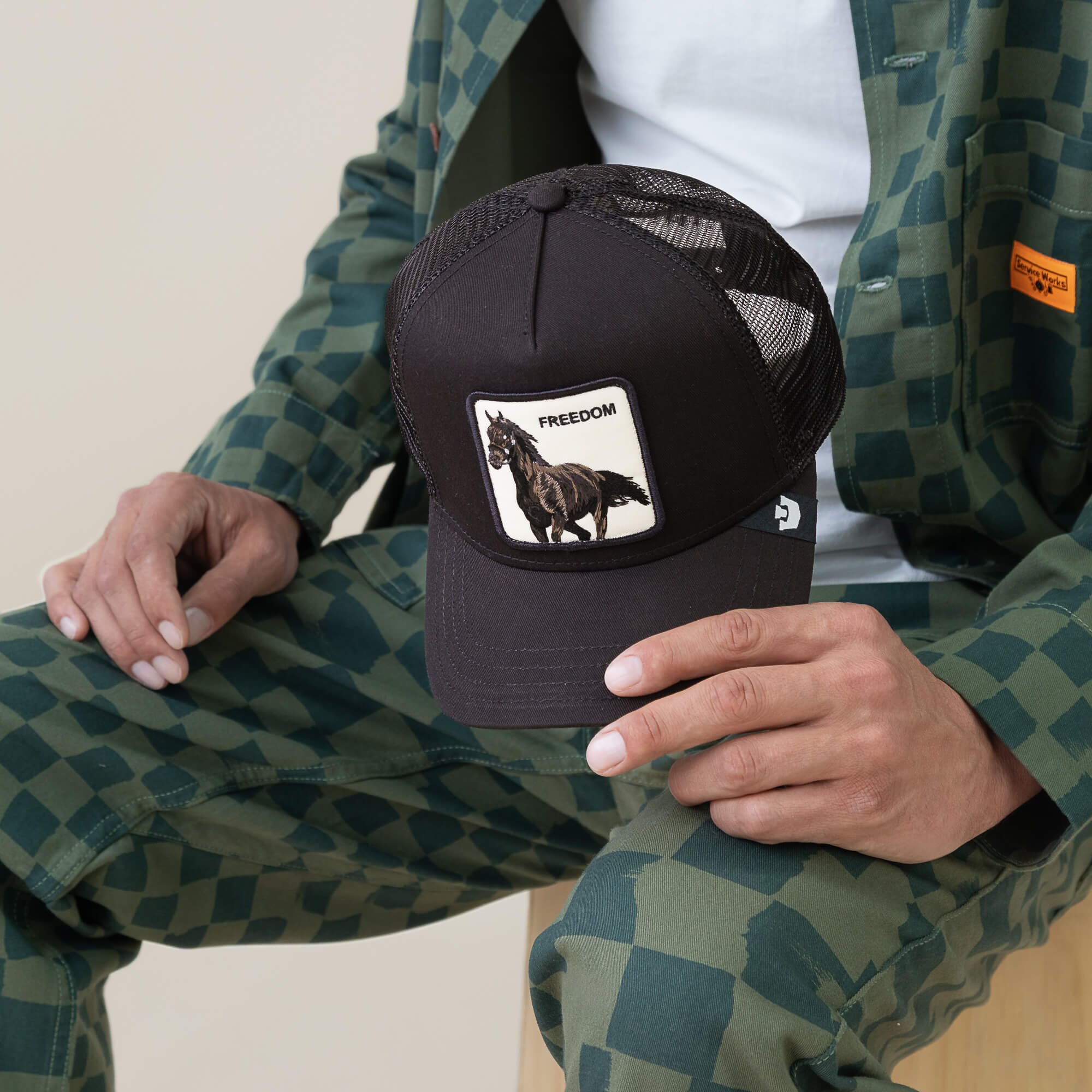 Wearing green checkered clothing, someone holds the Goorin Bros. Freedom Horse cap—a black mesh baseball hat with a running black horse patch and "FREEDOM," capturing wild energy and open-road spirit.