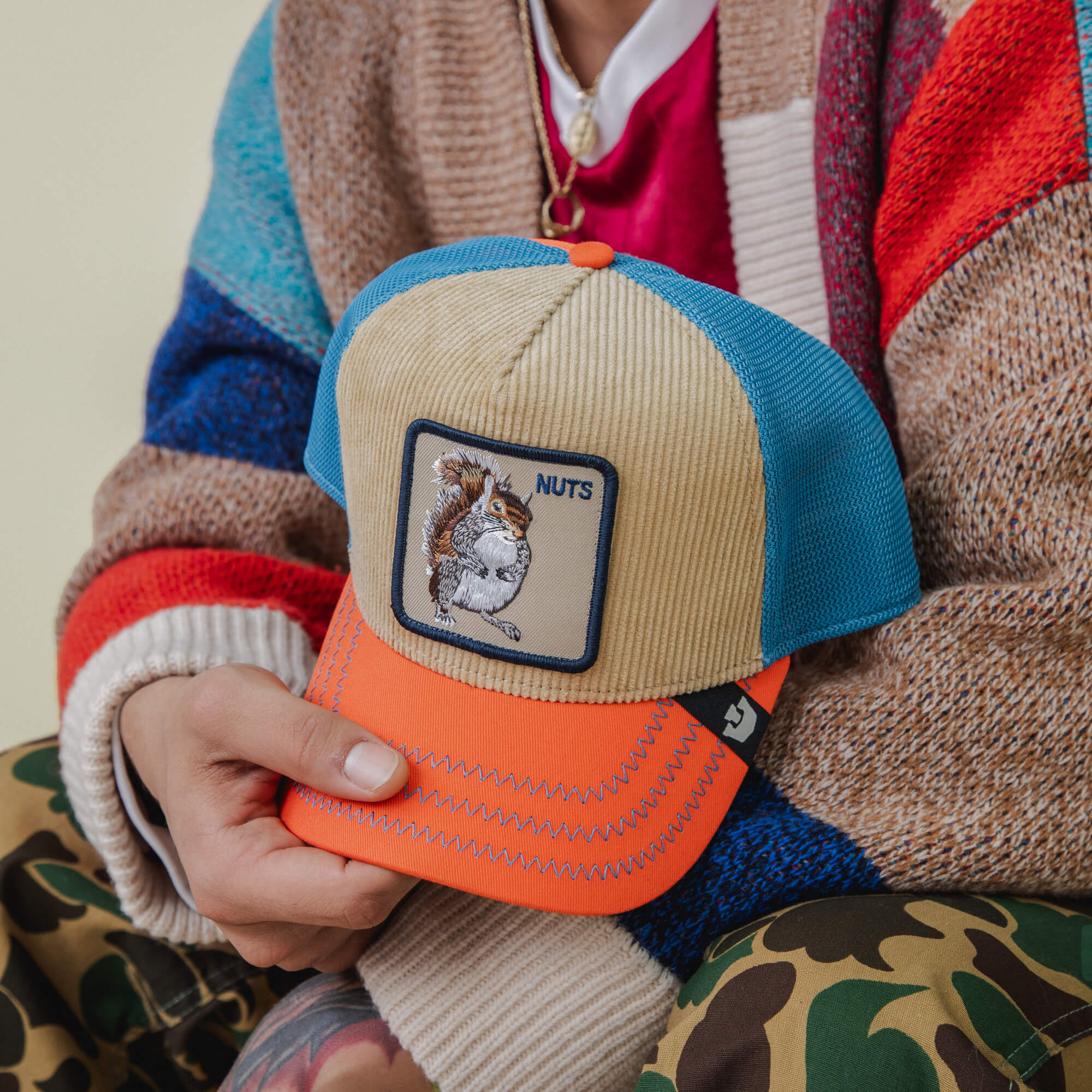 A person holds the Goorin Bros. Nuts Remix hat featuring a NUTS squirrel patch. Their electric blue mesh sweater pops against camo pants, giving off playful forest vibes.