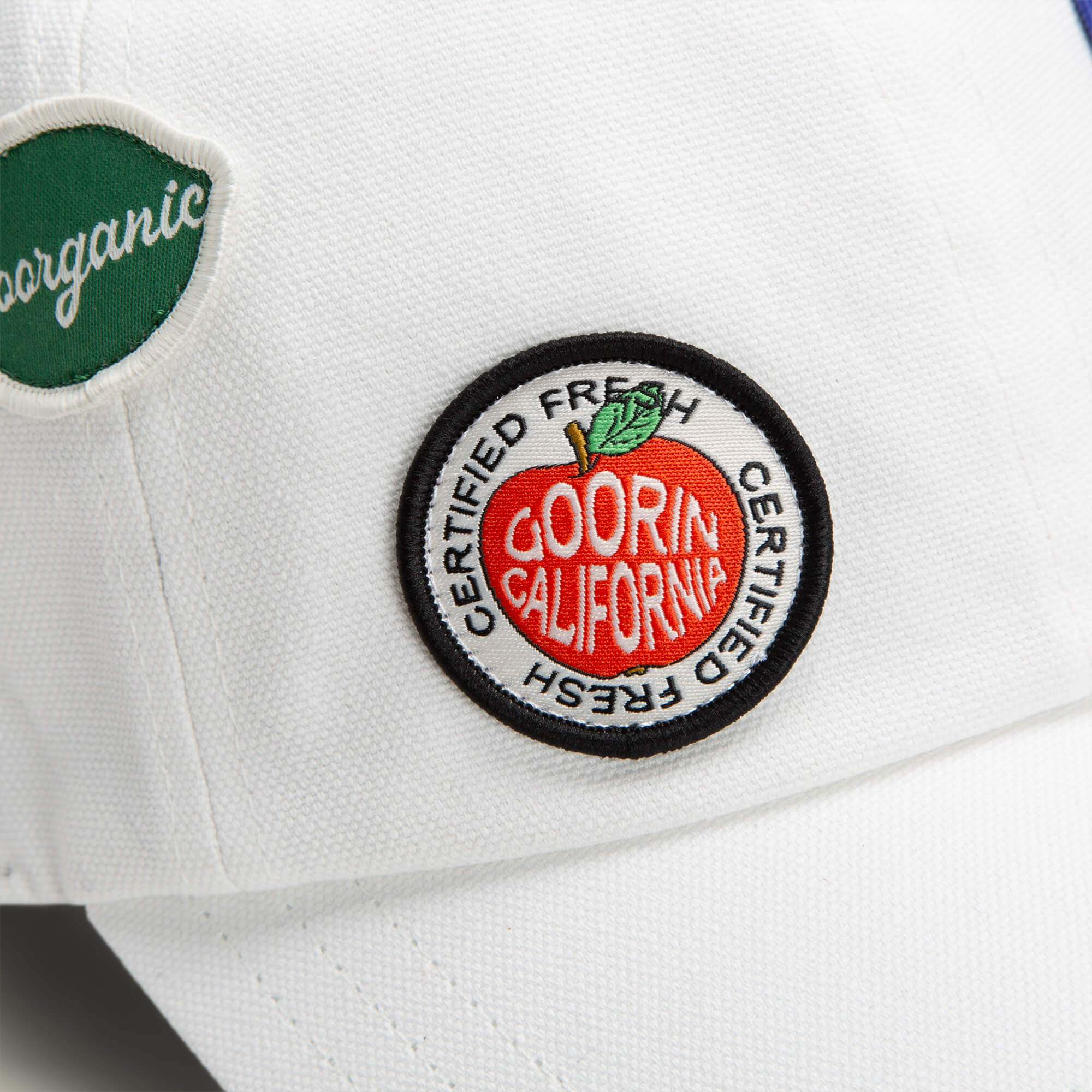 Close-up of Goorin Bros. Pop Appeal white cap with two patches: a green "organic" label and a circular patch showing a red apple with "Goorin California Certified Fresh" text.