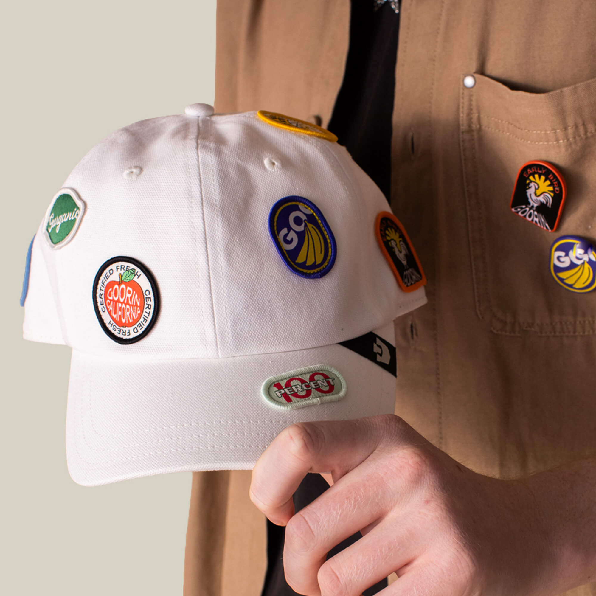 A person holds the Goorin Bros. Pop Appeal white canvas baseball cap featuring colorful embroidered patches, with similar badges on their tan shirt pocket.