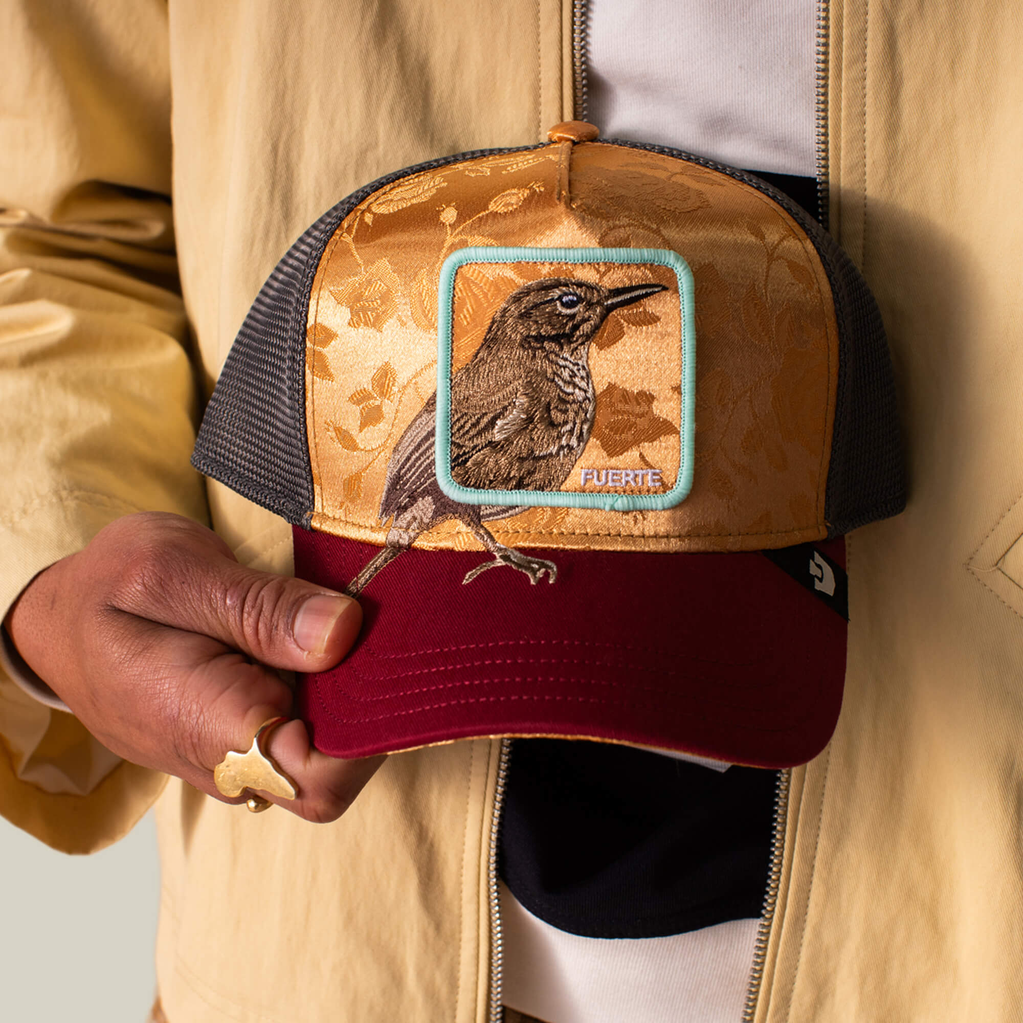A person holds the Goorin Bros. Costa Rica Thrush cap, featuring an embroidered thrush on the front. They wear a beige jacket and a gold animal-shaped ring, combining earthy tones with bold, nature-inspired style.
