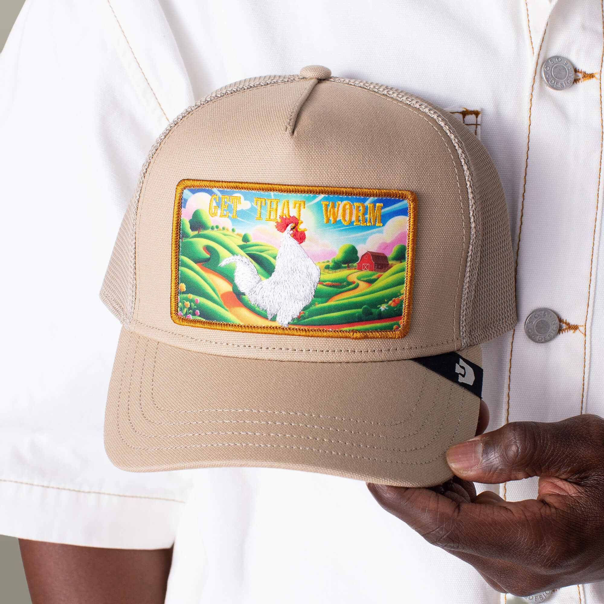 A person holds the Greener Grass - Get that Worm tan trucker hat by Goorin Bros., featuring a colorful rooster patch and "GET THAT WORM" text, against a scenic farm backdrop.
