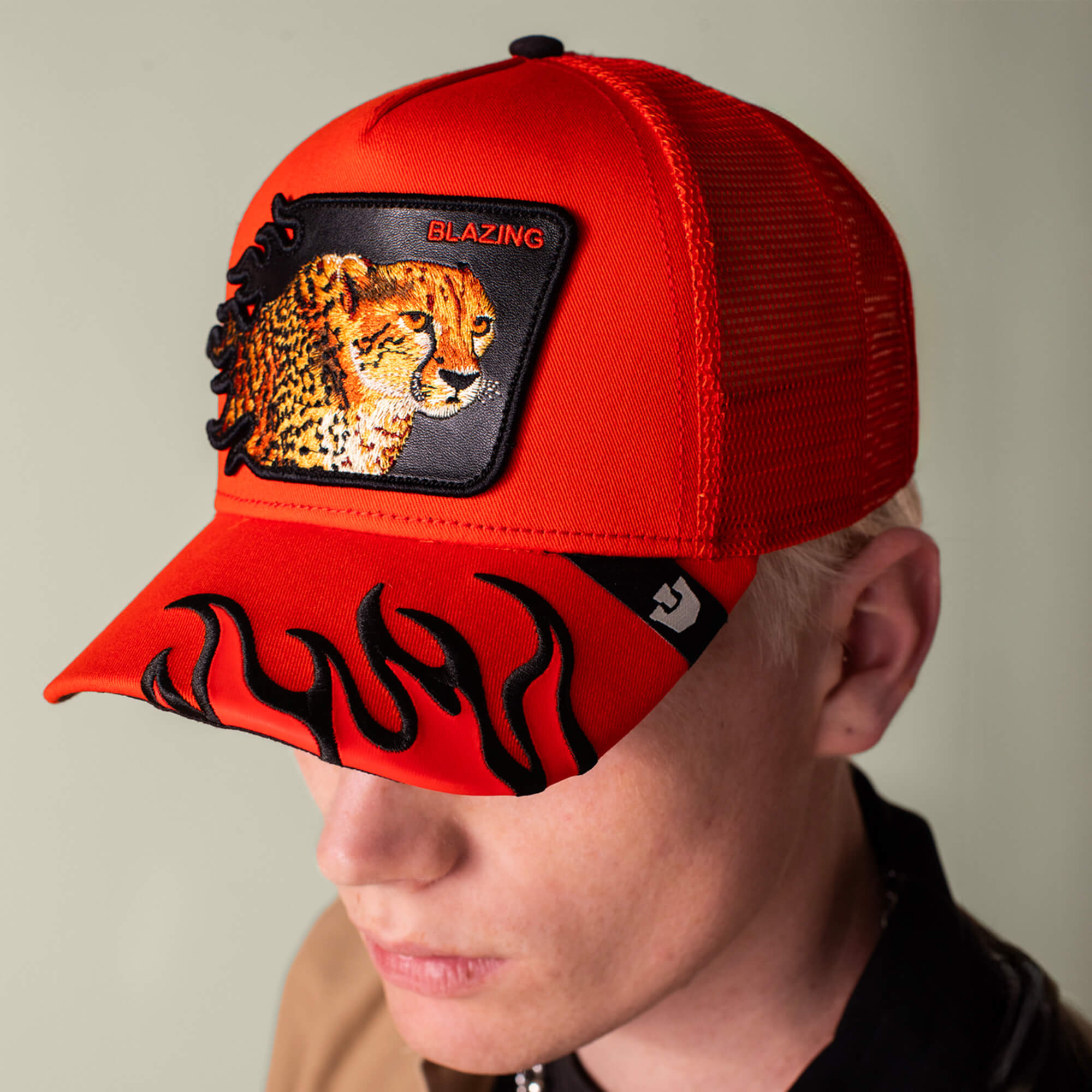 A person wearing a vibrant red mesh Goorin Bros. Blazing Flames trucker cap with a cheetah patch marked "BLAZING" and bold flame accents on the brim.
