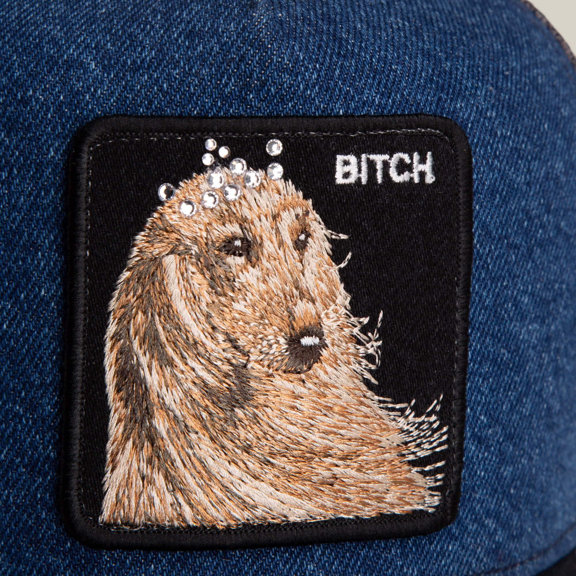 The Hound Dawg patch by Goorin Bros. features an embroidered hound dog with a jeweled denim crown and “BITCH” lettering on blue denim fabric.