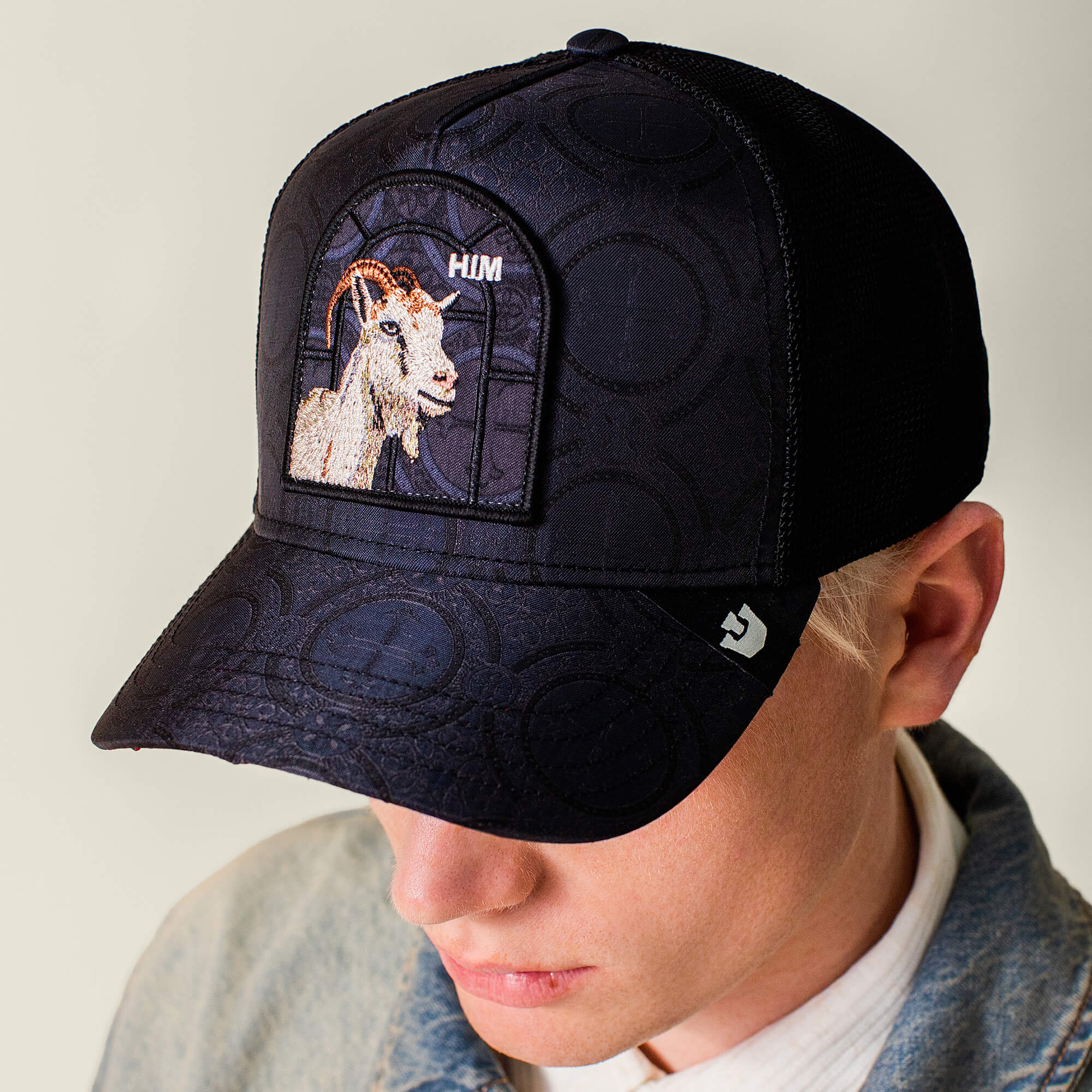 A person wearing a Goorin Bros. Religulous - Hymn charcoal black hat with an embroidered goat and "HUM" on the front, paired with a light denim jacket and white shirt.