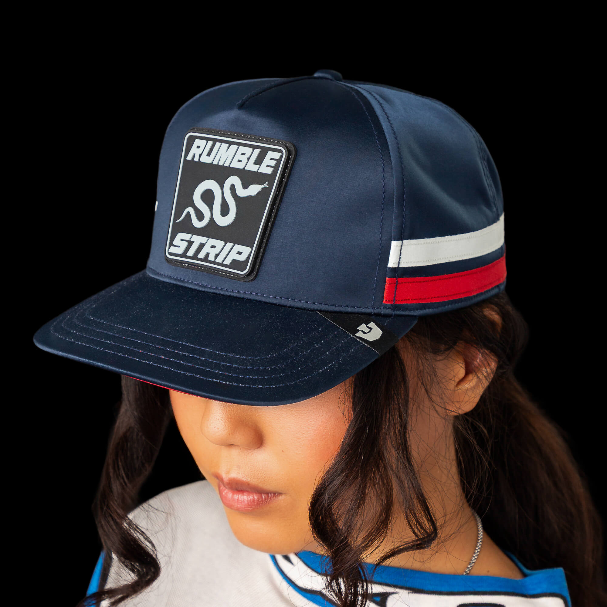 A person in a Goorin Bros. Pole Position - Rumble Strip cap, featuring red, white, and blue stripes and a signature patch, looks downward against a black background—a style popular among drift drivers.
