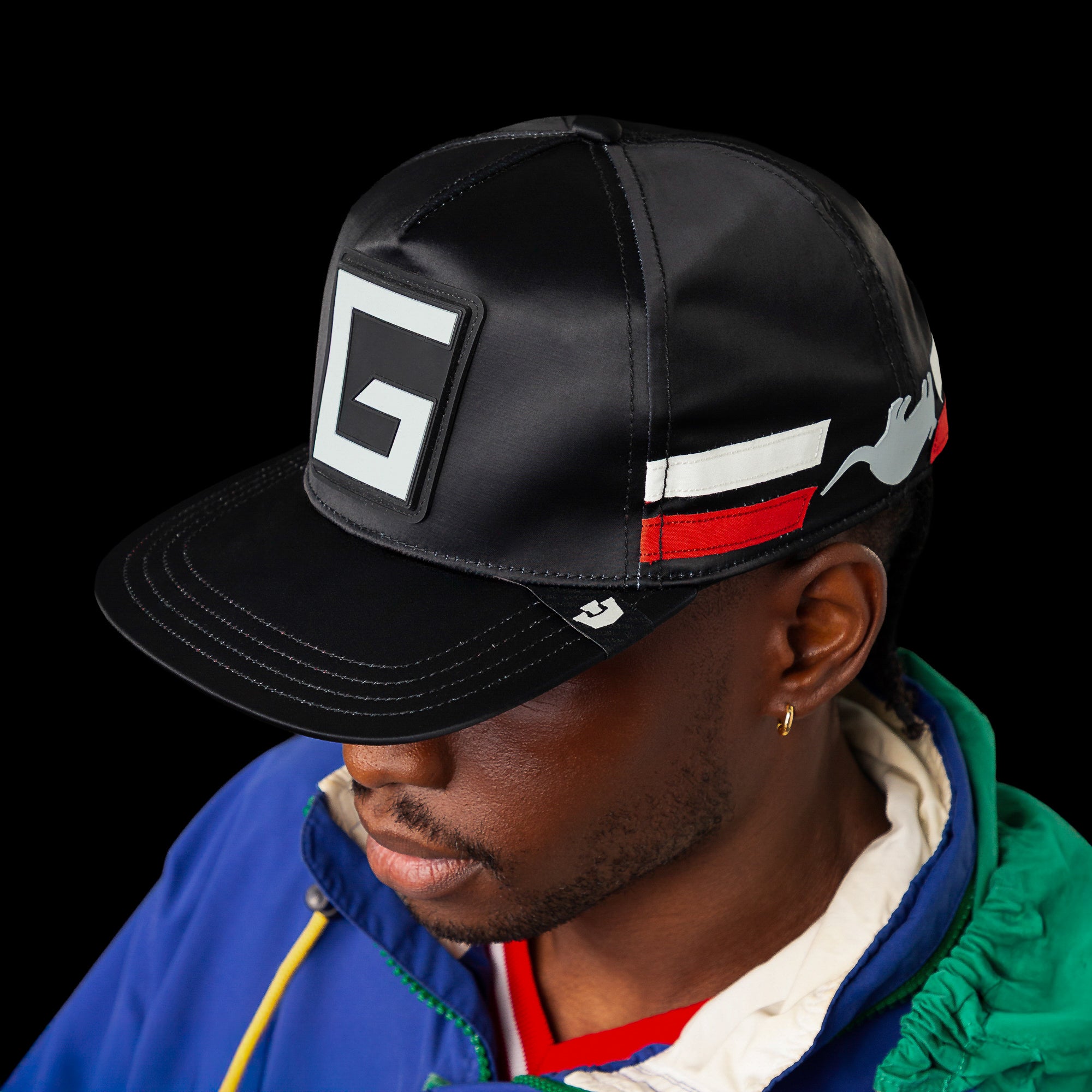 Man wearing a black Goorin Bros. Pole Position - Road Kill hat with a large G on the front, red, white, and gray side stripes, paired with a blue jacket featuring a white hood.