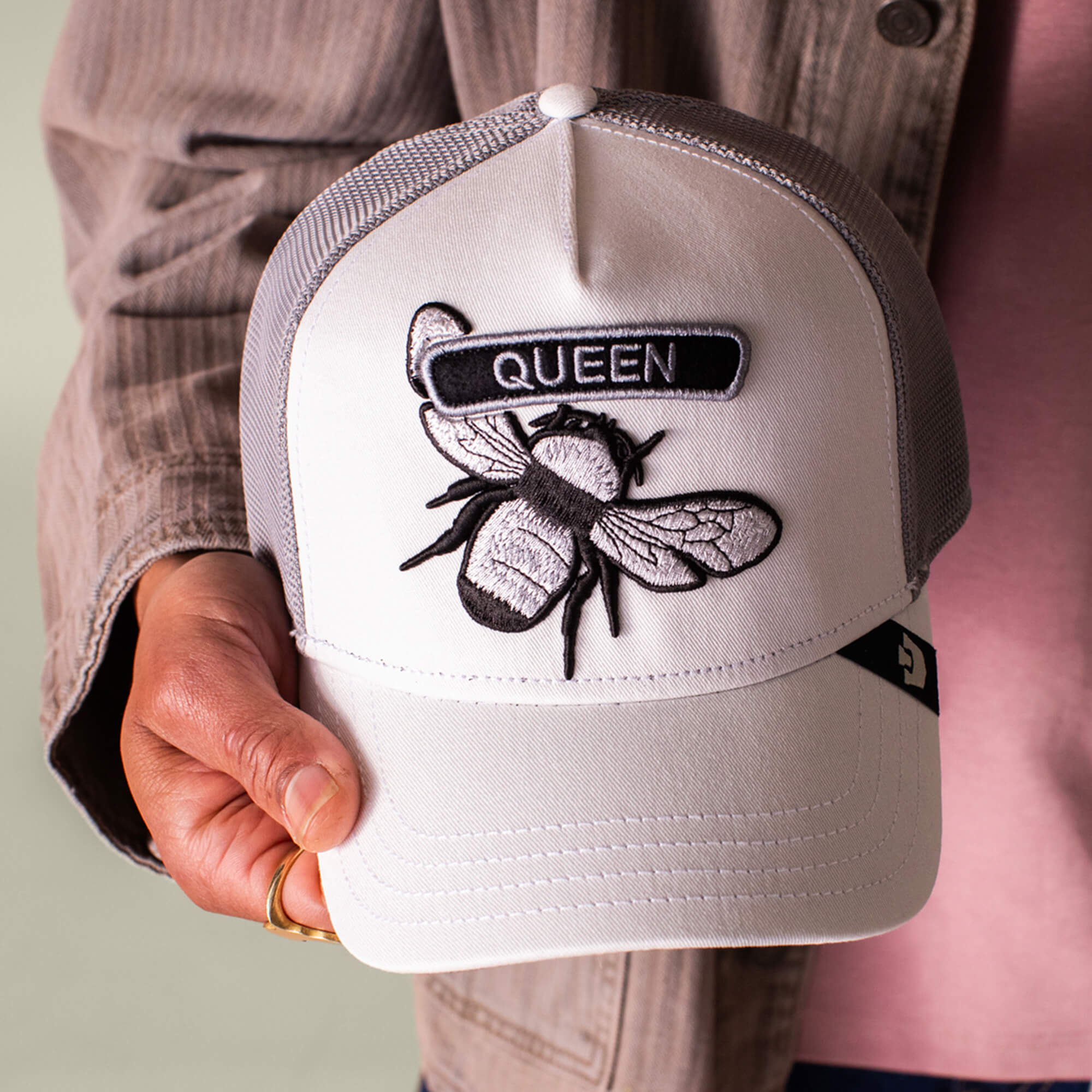 A person holds the Goorin Bros. Classic Rocker - Queen hat, a white and gray mesh cap featuring an embroidered bee patch with "QUEEN" above it, capturing iconic rocker Queen style.