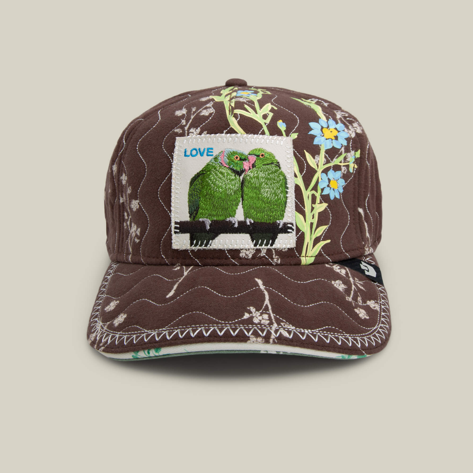 Chocolate brown quilting and hand-stitched details accent the Goorin Bros. Vol. 1 - Quiltbag: Love trucker cap in OXIDE, with white floral stitching and a patch showing two green parrots under the word LOVE.