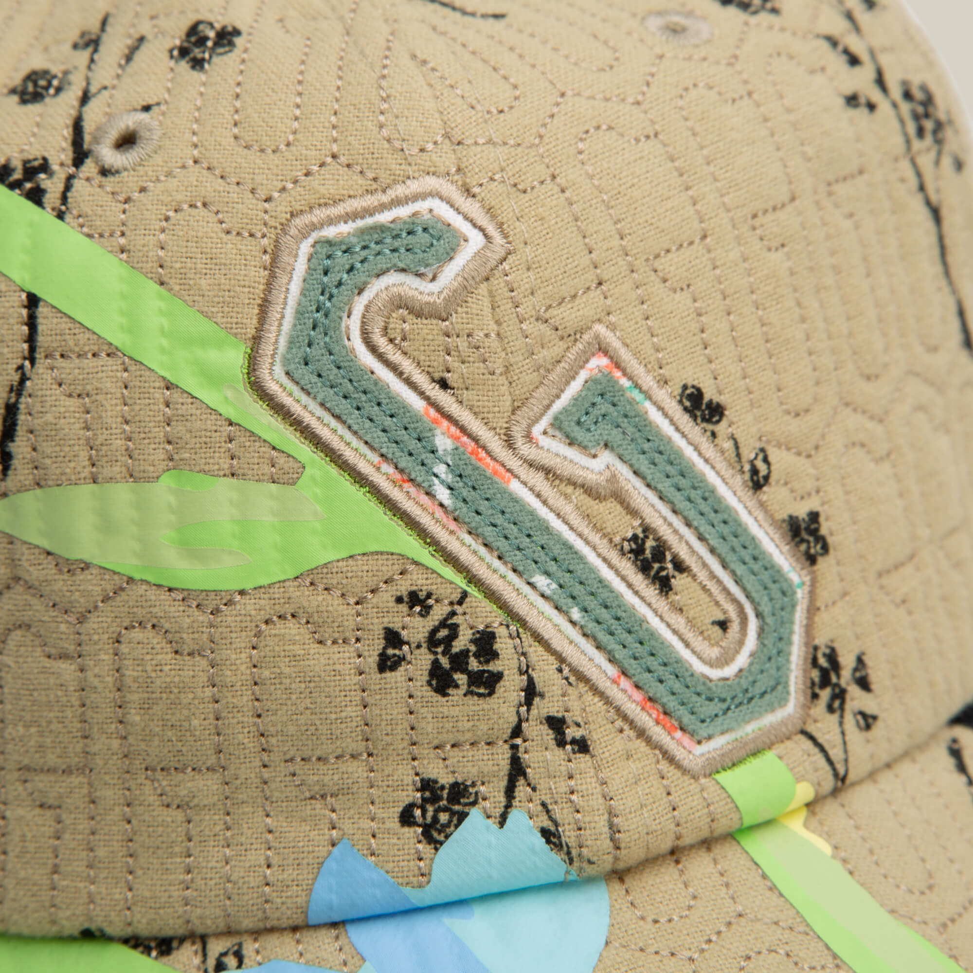 Close-up of the Vol. 1 - Quiltbag: G Papa cap by Goorin Bros.—a beige quilted papa cap featuring a green and white embroidered letter G, black floral patterns, and green and blue abstract shapes.