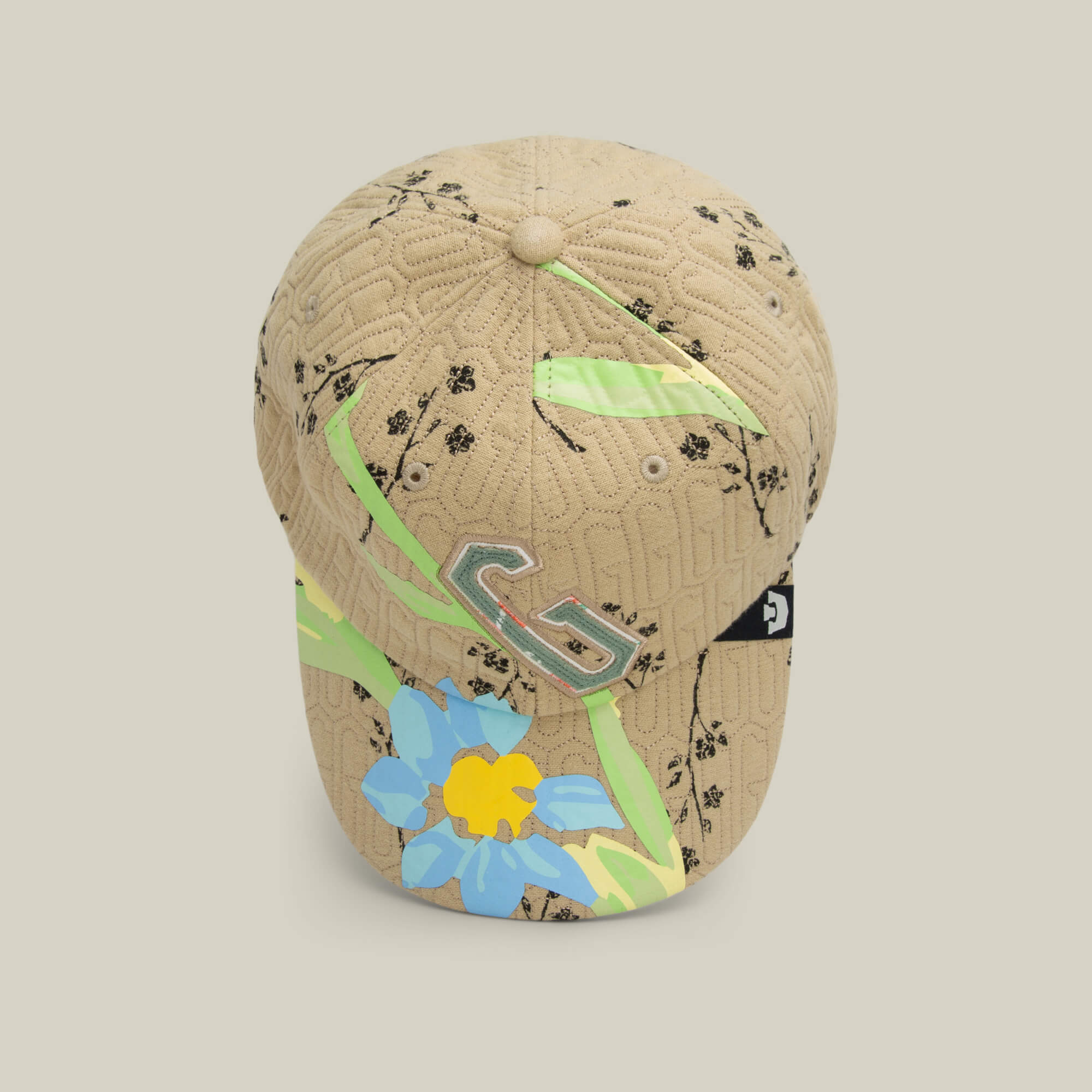 The Vol. 1 - Quiltbag: G Papa cap by Goorin Bros. features beige quilted floral fabric with black patterns, green and yellow accents, a striking blue flower, and a metallic "G" logo for standout style.
