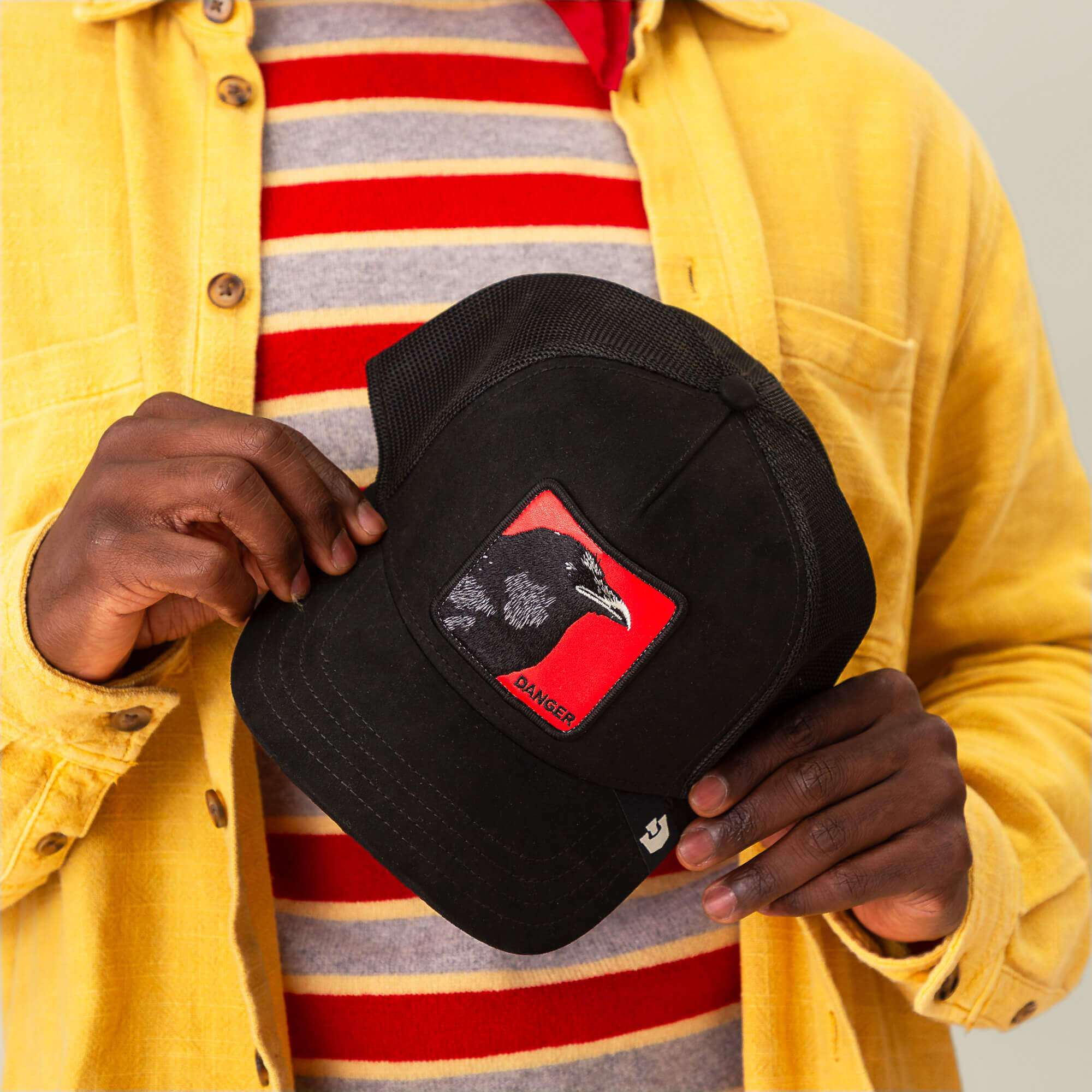 A person in a yellow shirt holds a black Goorin Bros. Corvo Rosso cap featuring a patch with a sharp-eyed bird and "DANGER" on a signal red background, showcasing the bold Corvo Rosso style.