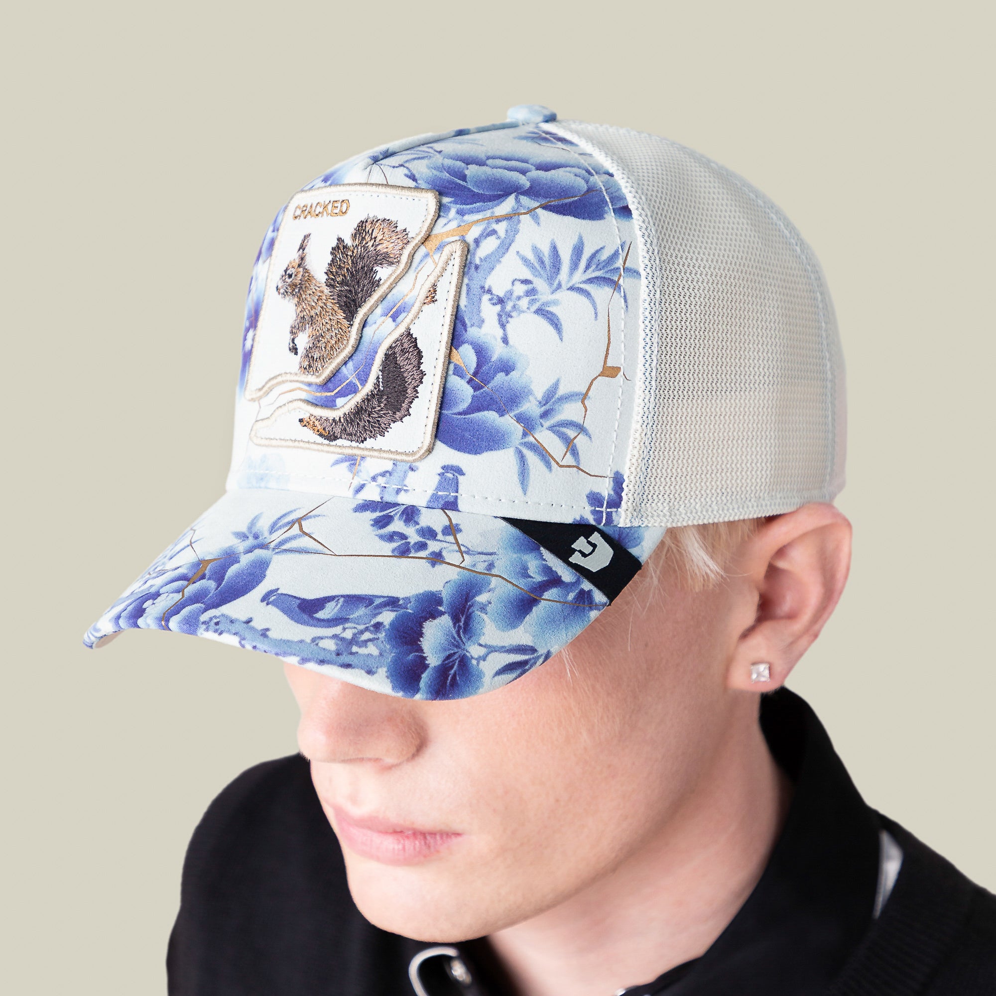 A person wears the Goorin Bros. "Break The Mold" white mesh baseball cap featuring blue floral designs and a squirrel patch, paired with a black shirt and a stud earring in their left ear to express their unique style.