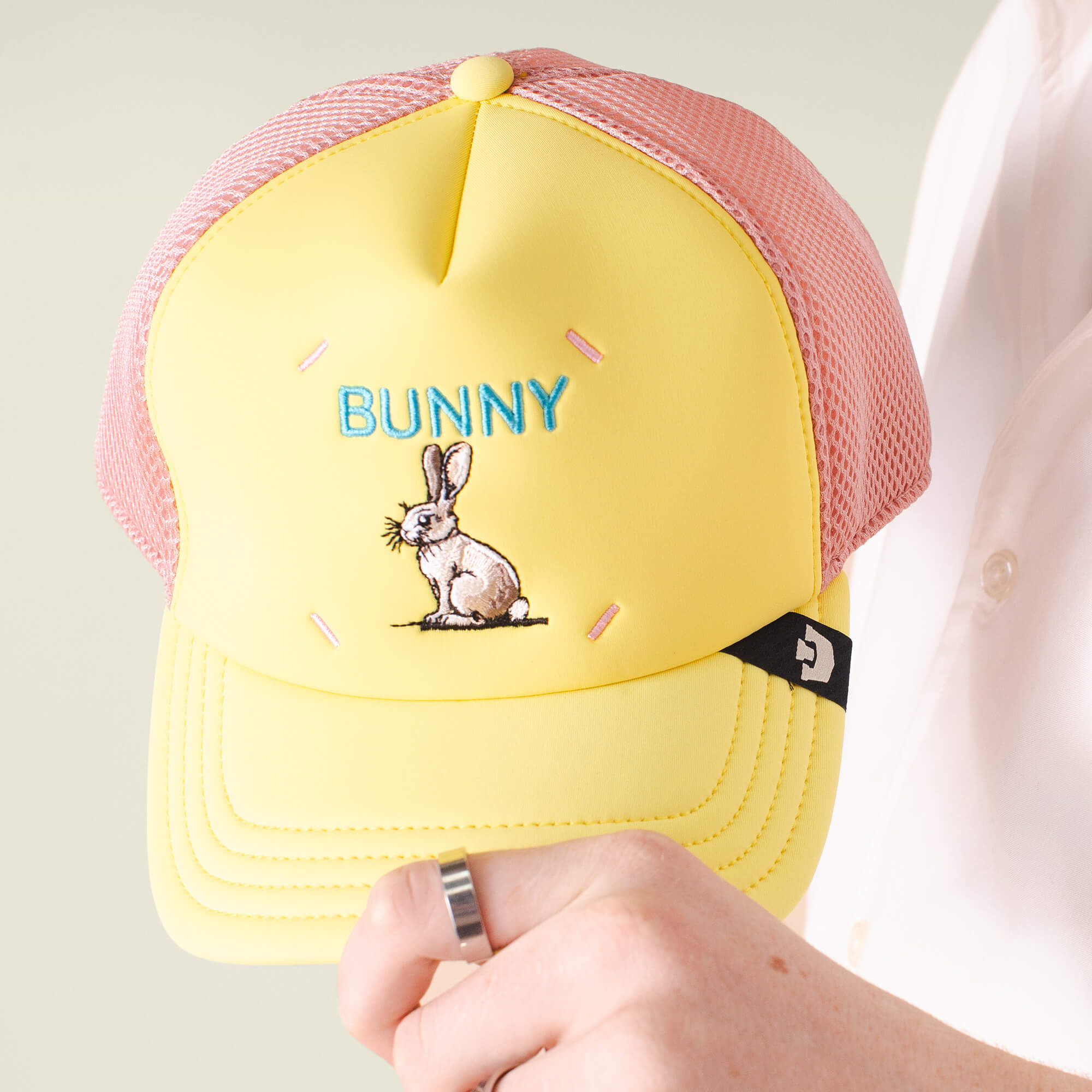 A hand holds the Goorin Bros. Neo Farm - Bunny Rabbit hat, featuring a playful yellow and pink mesh design, an embroidered bunny and "BUNNY" in blue—perfect for everyday wear.