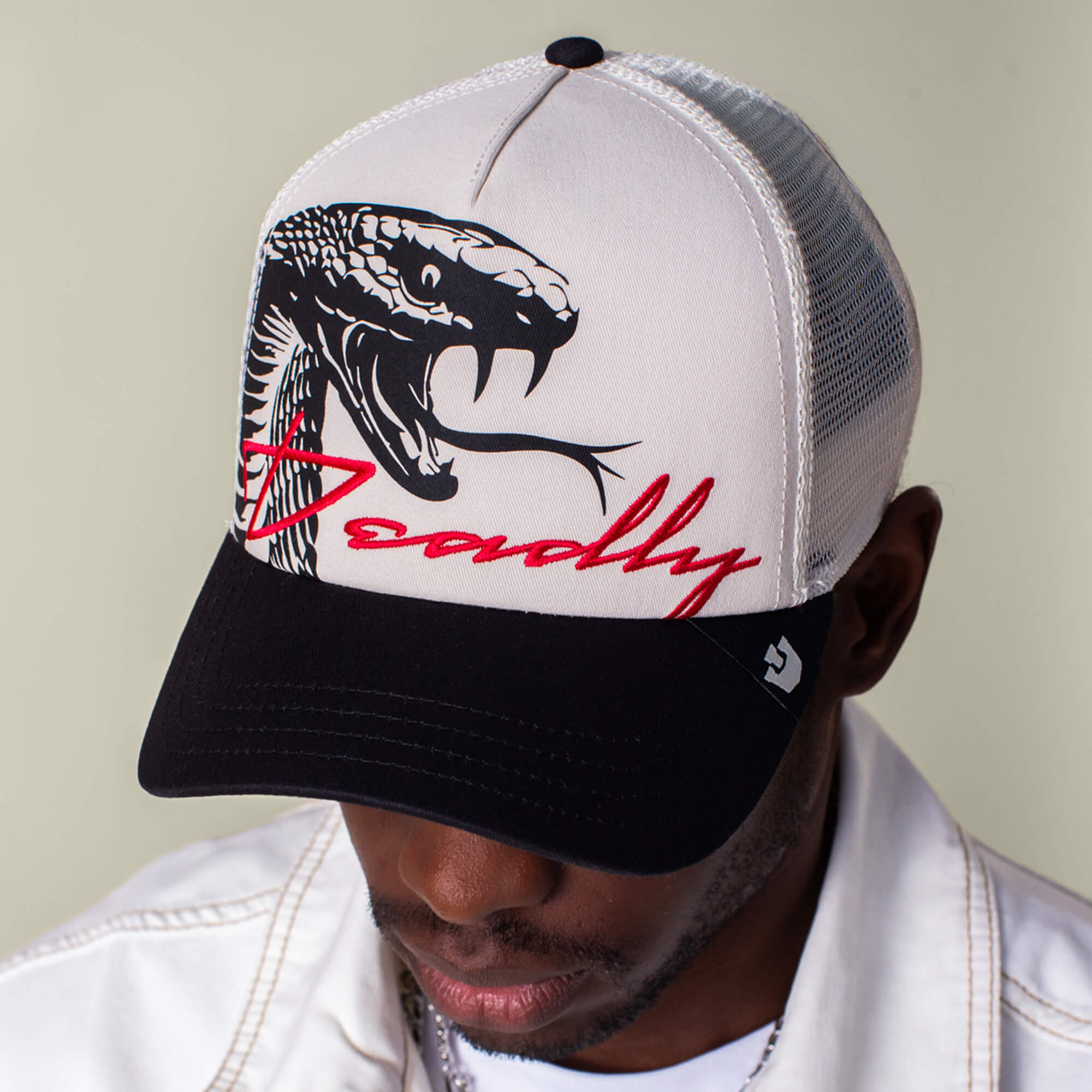 Man wearing a Goorin Bros. "Outside the Lines Snake" cream and black patch hat with bold red "Deadly" script, paired with a white jacket. His face is partially obscured by the hat's red script.