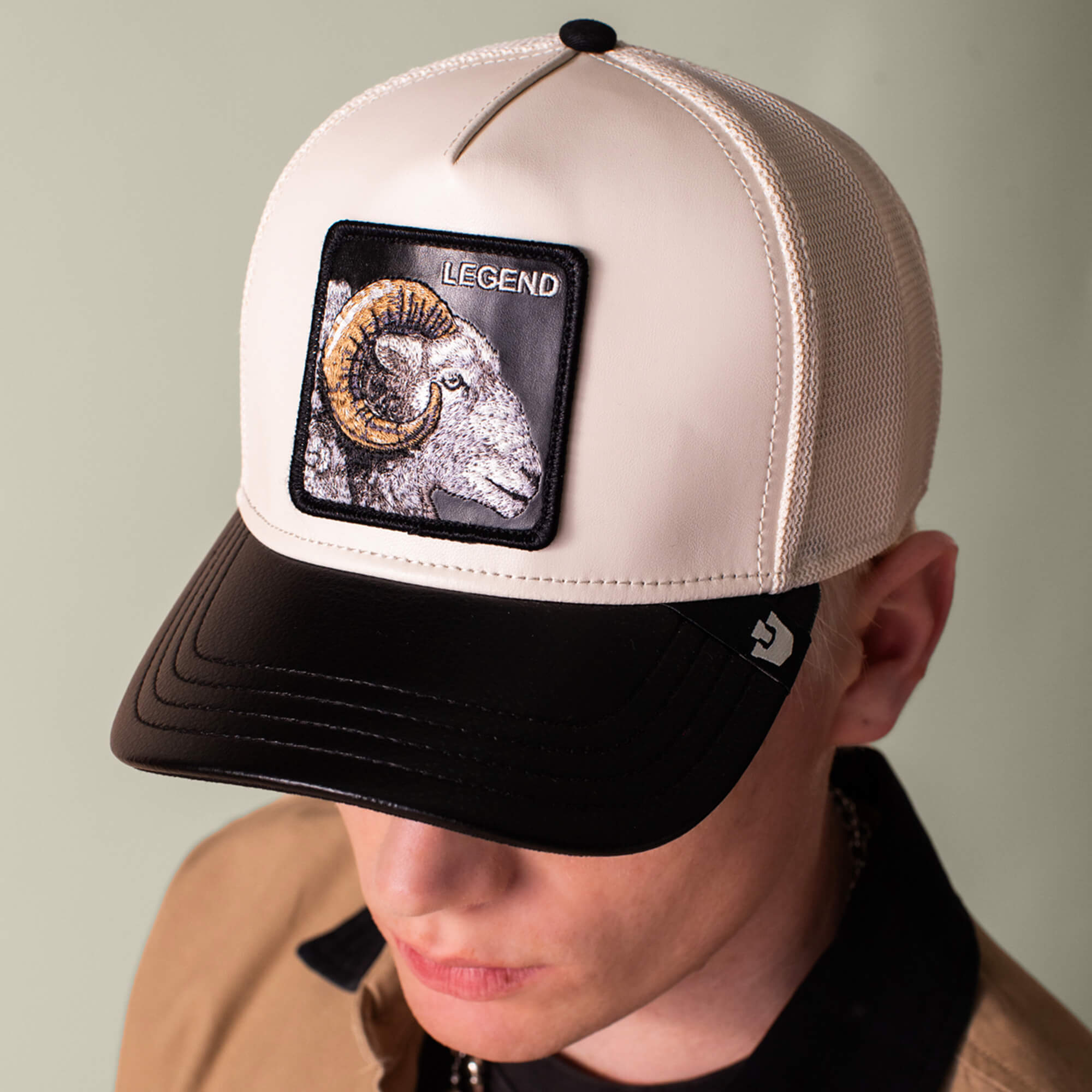 A person wears the Goorin Bros. Faux Leather Ram cap, featuring a two-tone beige and black design with a LEGEND patch on the front. The neutral cream colorway adds versatility to this stylish faux leather hat.