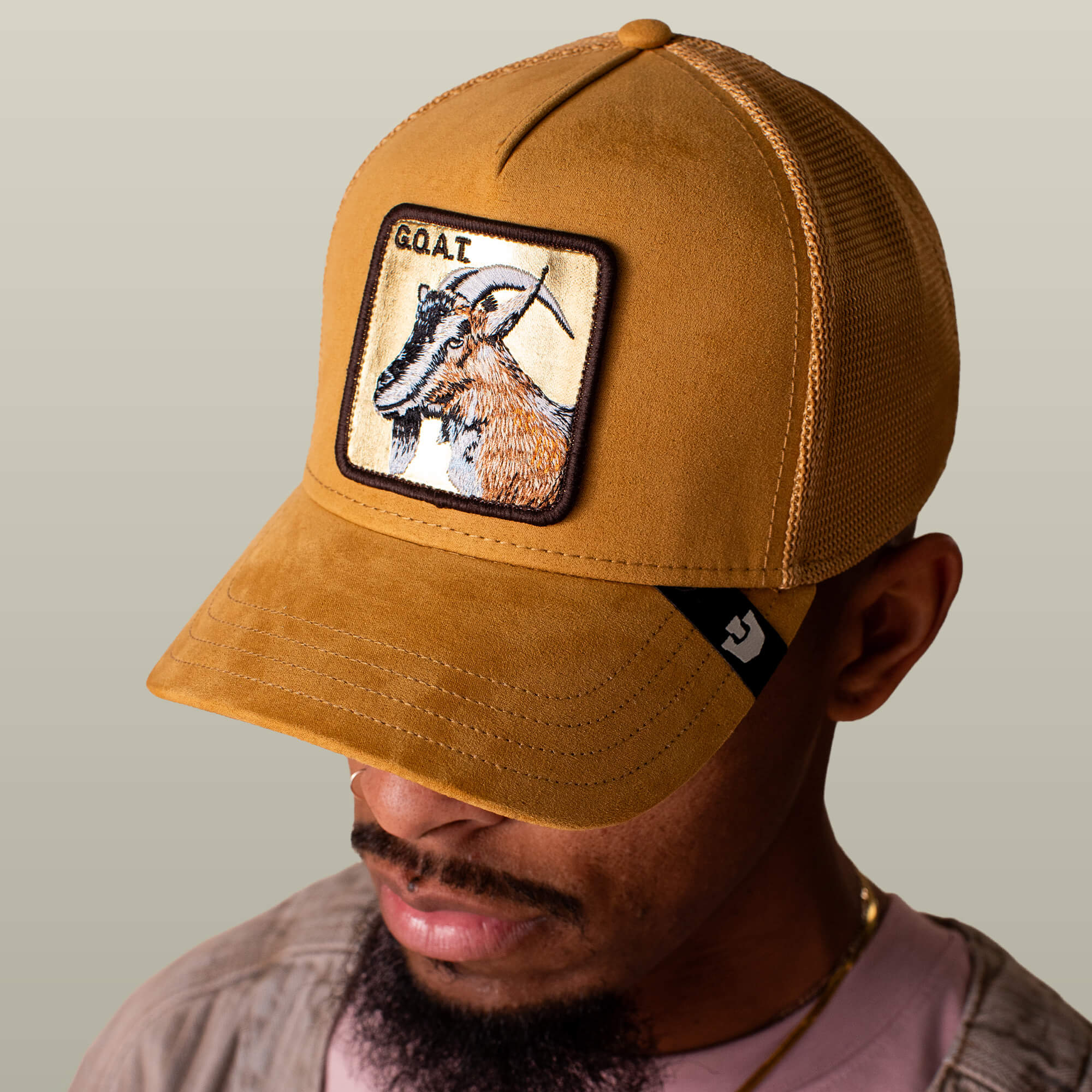 A man wears the Goorin Bros. Golden Goat tan gold suede trucker hat featuring a goat patch and "GOAT" text on the front, set against a plain neutral background that highlights his confident style.