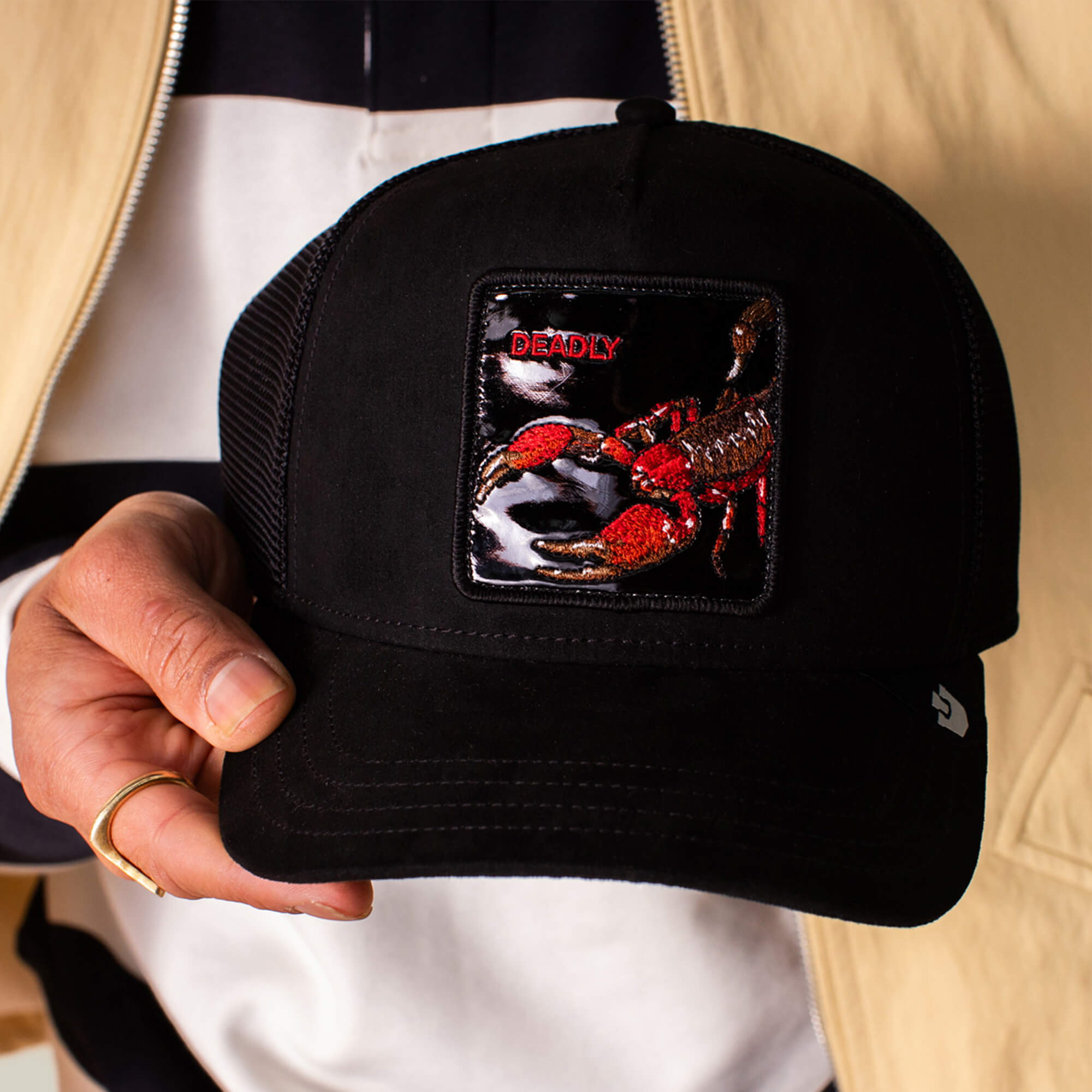 A person holds the Goorin Bros. Black Out Scorpion cap, a sleek black suede baseball hat featuring a scorpion patch with "DEADLY" embroidered above.
