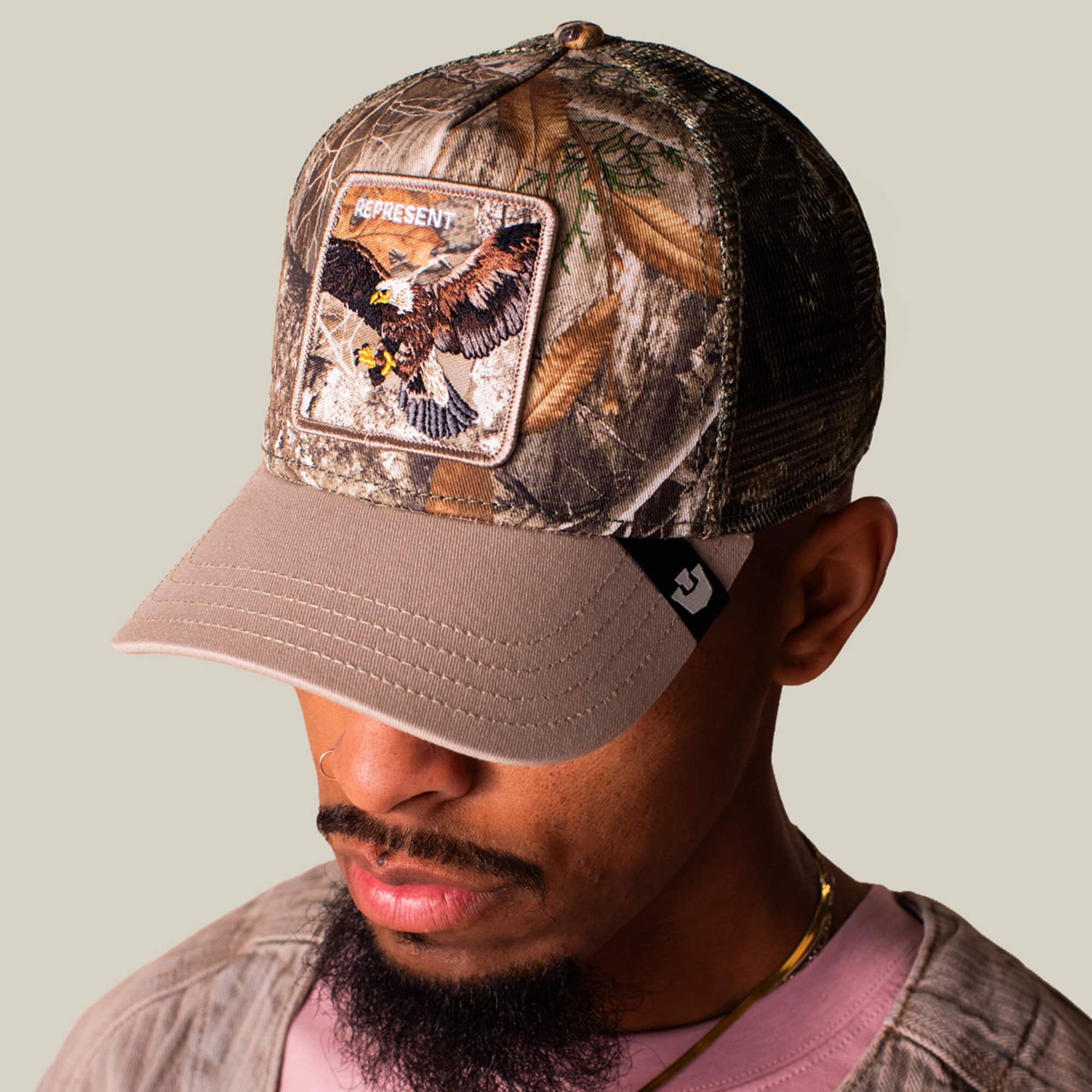 A man wears the Goorin Bros. Realtree Edge Represent Eagle hat, featuring camo print, an embroidered eagle patch, and “REPRESENT” on the front, as he looks down against a plain background.