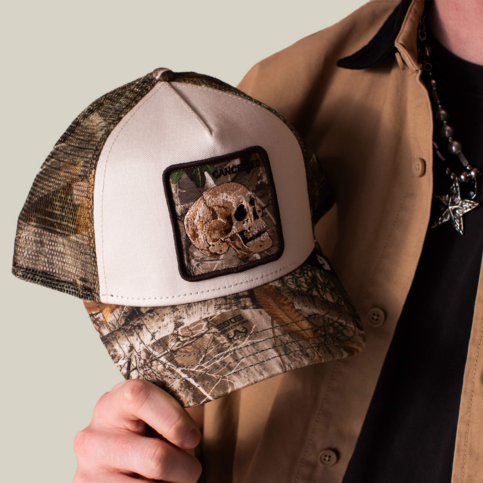 A person holds the Goorin Bros. Realtree Edge Cancelled Skull baseball cap with a skull patch; they wear a tan shirt, black shirt, and a beaded necklace with a star pendant.
