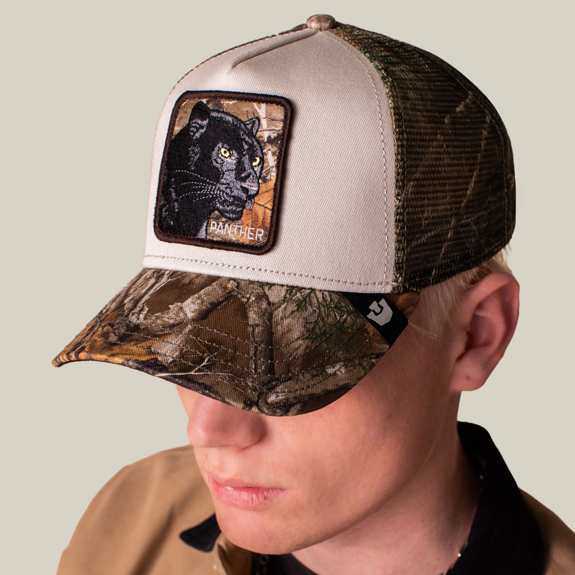 A person wears the Goorin Bros. Realtree Edge Black Panther trucker hat in beige and camo, featuring a bold panther patch on the front, looking downward.