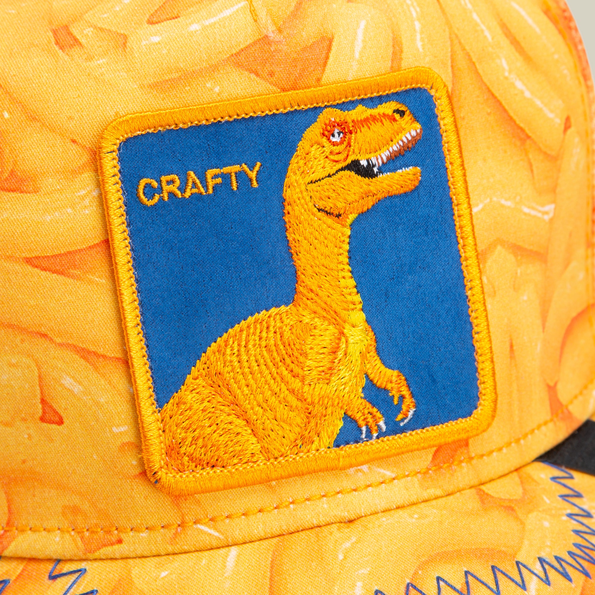 A yellow trucker hat with a patch showing a yellow dinosaur on a blue background and the word CRAFTY in yellow text. Featuring bold hat designs, this Goorin cap has a yellow pattern and visible blue stitching.
