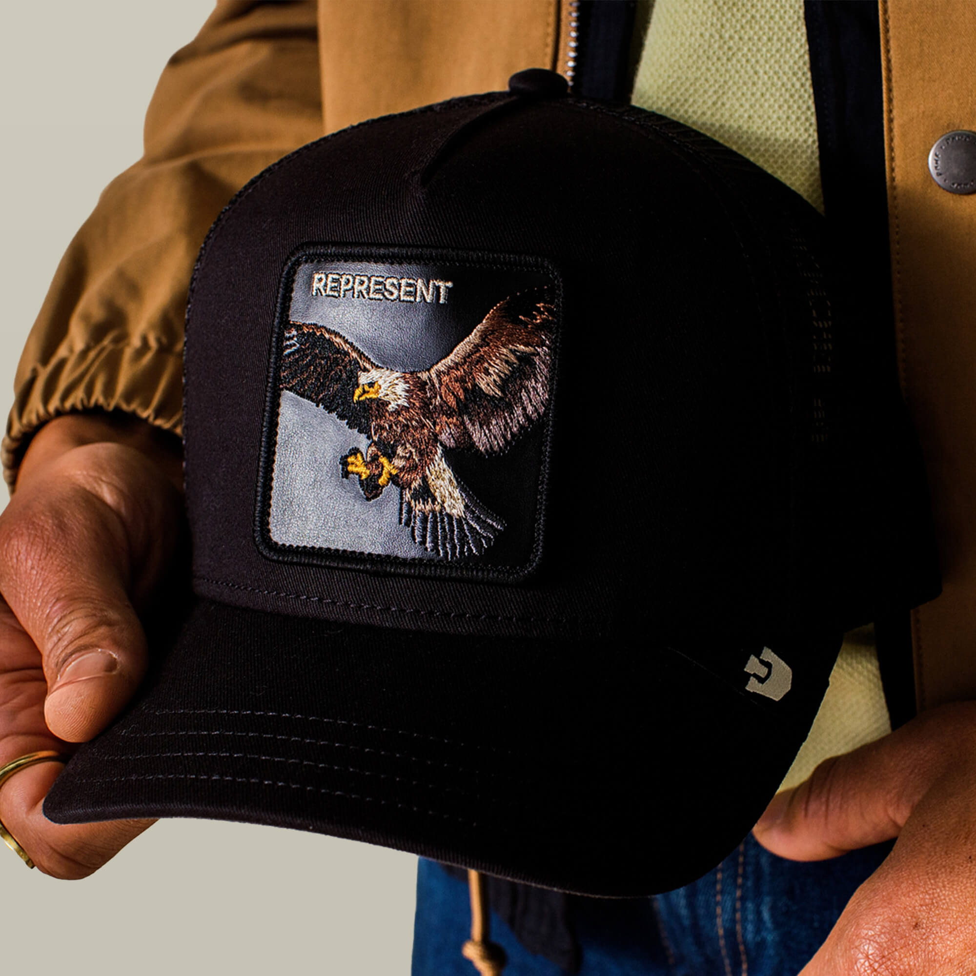 A person holds the Goorin Bros. Represent Eagle Trucker hat, which features an embroidered eagle patch with "REPRESENT" stitched above it.