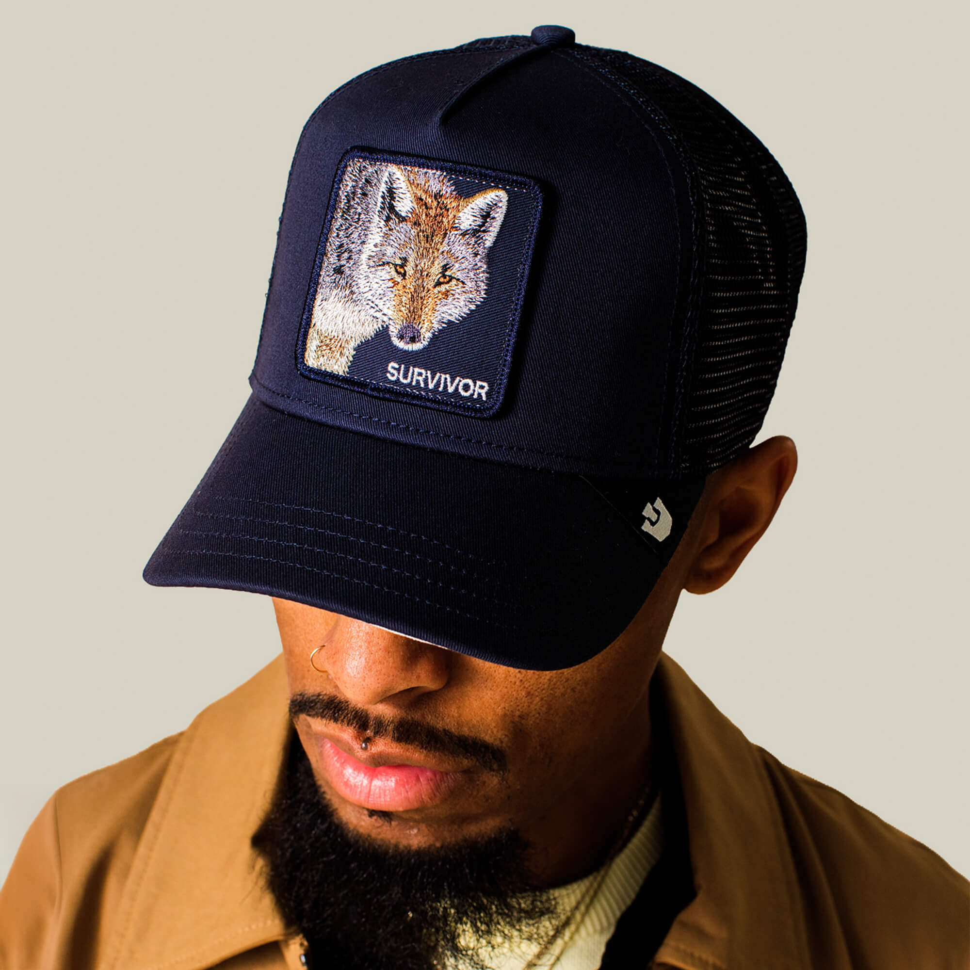 Man wearing the Goorin Bros. Mirage Survivor trucker hat featuring a fox patch labeled SURVIVOR, paired with a tan jacket and beige shirt, looking down.