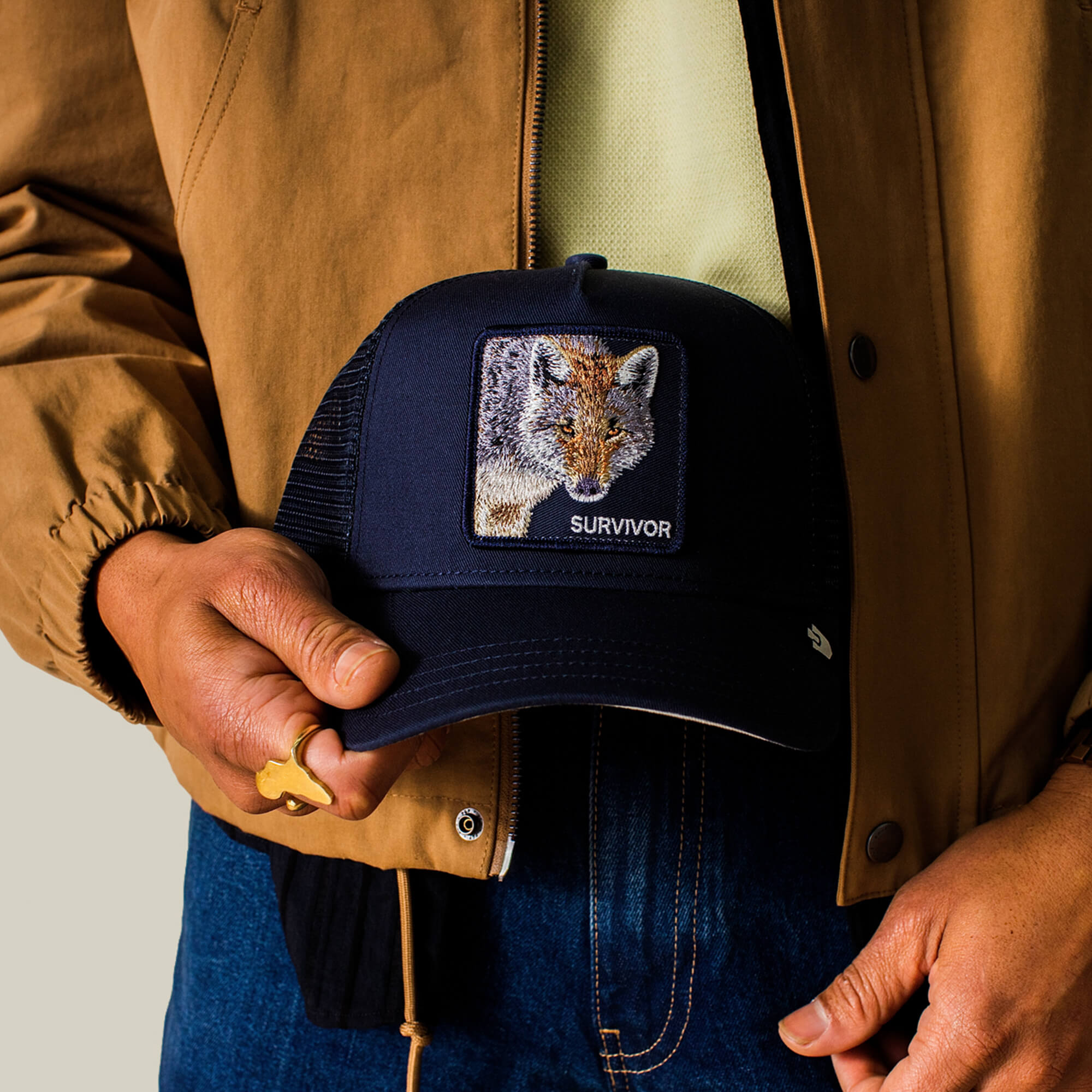 A person in a brown jacket holds a navy Goorin Bros. Mirage Survivor trucker hat featuring a fox patch labeled "SURVIVOR.