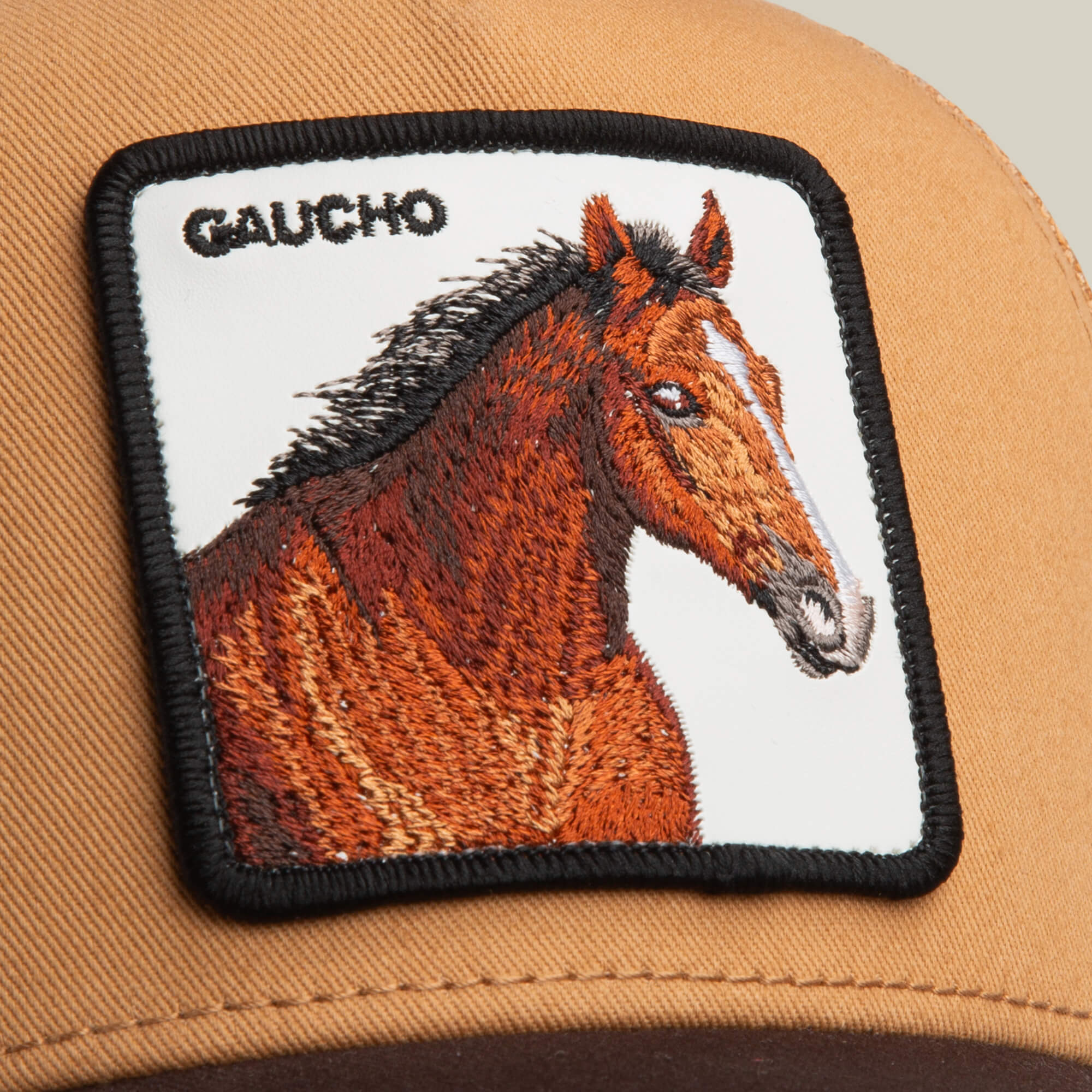 Close-up of the Gaucho Trucker by Goorin Bros., a tan earthy cap featuring a square horse patch with detailed horse art, leather accents, and GAUCHO embroidered in black.