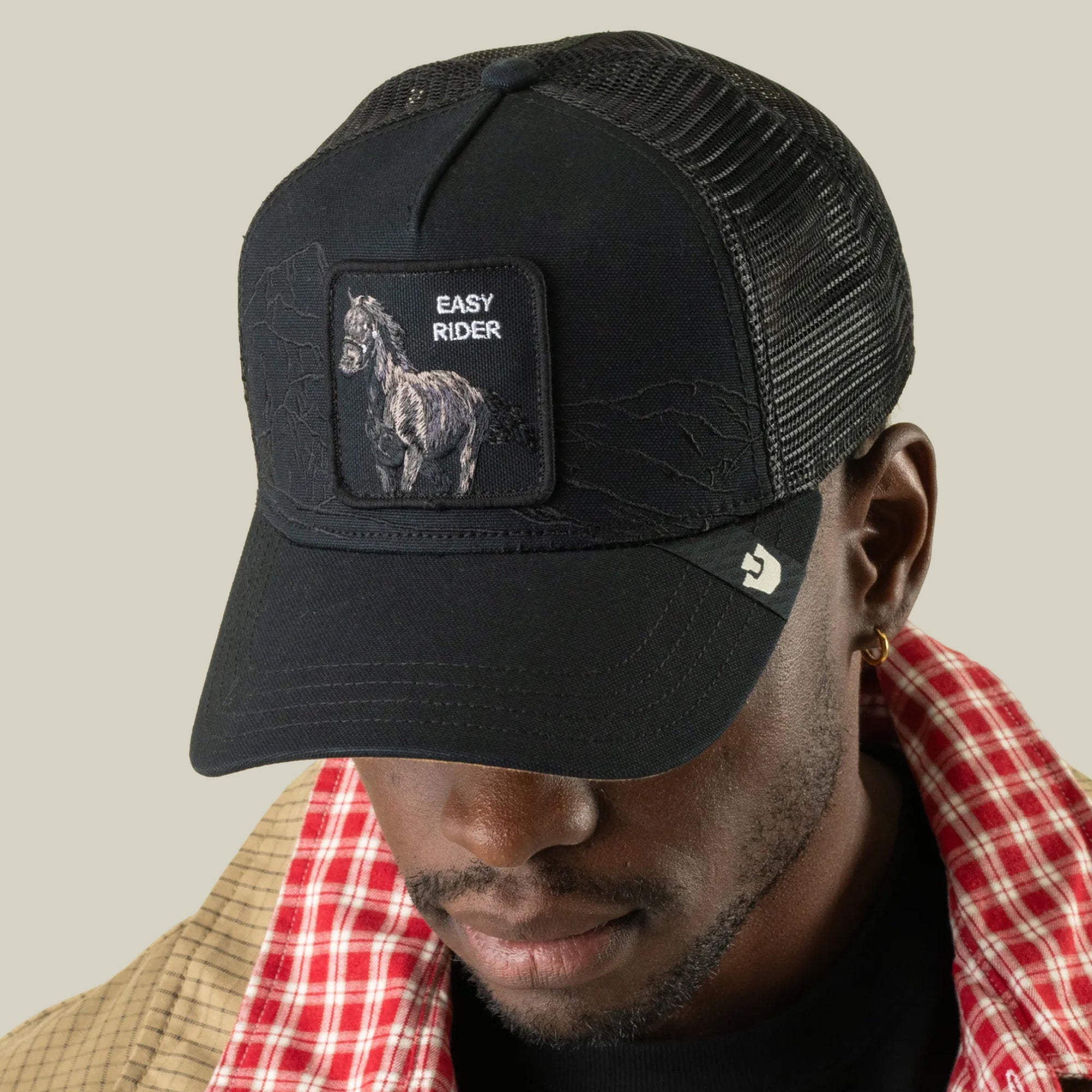 A man wears the Goorin Bros. Badlands Stallion—a black trucker hat featuring breathable mesh and a horse patch labeled EASY RIDER, enhanced by notable badlands stitching that adds subtle detail to this classic cap. He is paired with a plaid jacket and looks downward against a plain background.