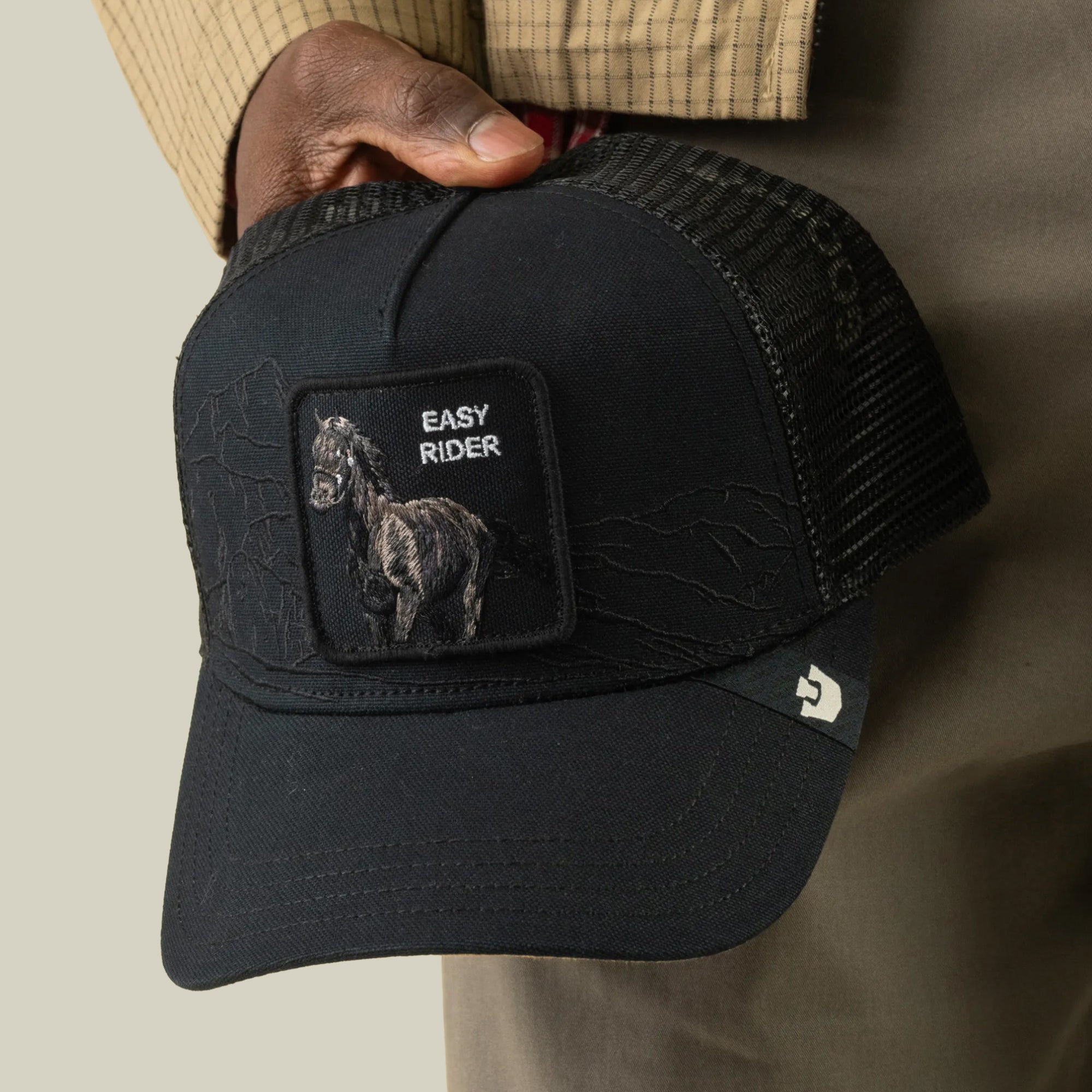 A person holds a black Goorin Bros. “Badlands Stallion” trucker hat with breathable mesh and a patch featuring a horse and the text EASY RIDER on the front.