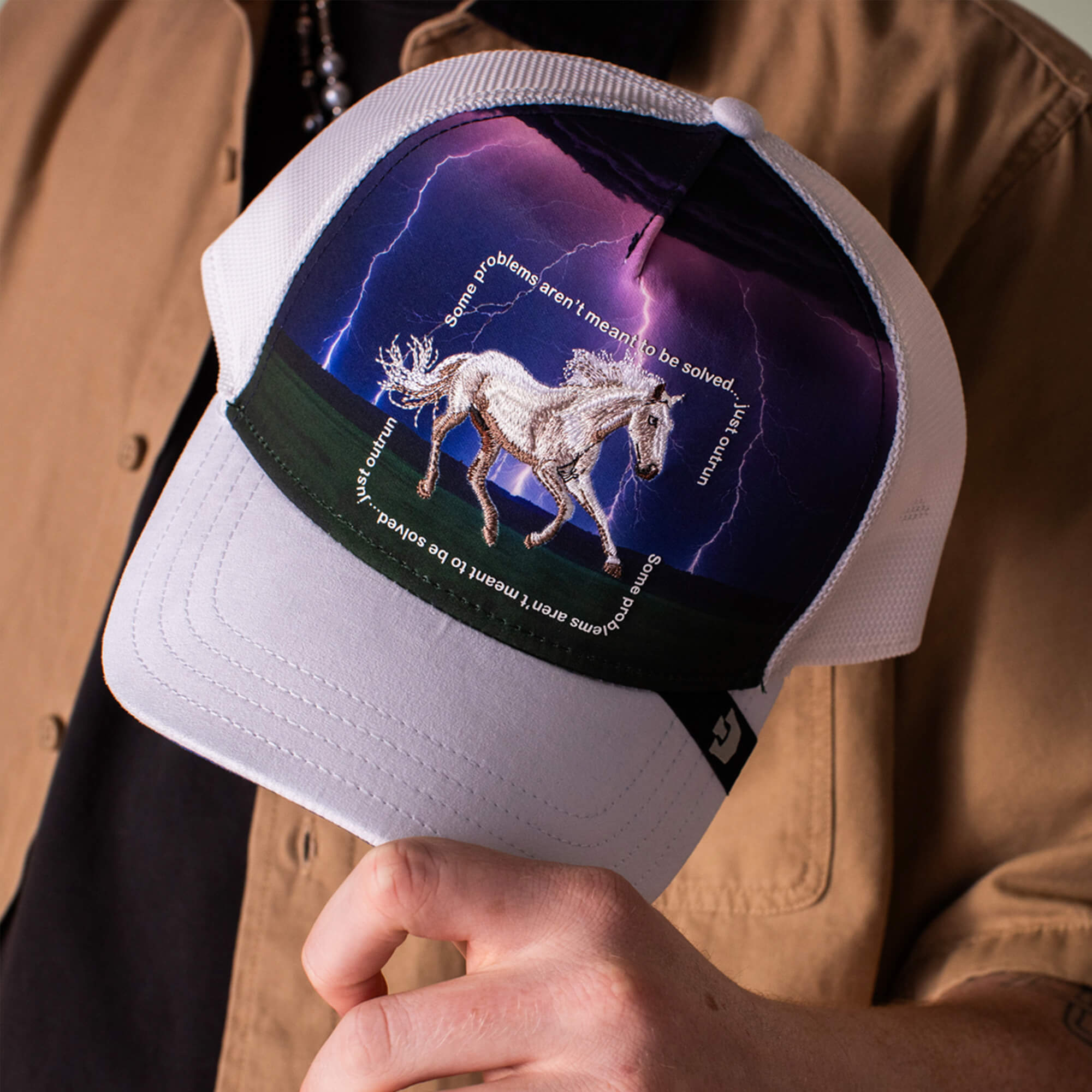 Someone holds the "Through the Storm - Horse" white trucker hat by Goorin Bros., featuring a horse graphic, lightning, and the phrase “Some problems aren’t meant to be solved. Run, unicorn, run.”.