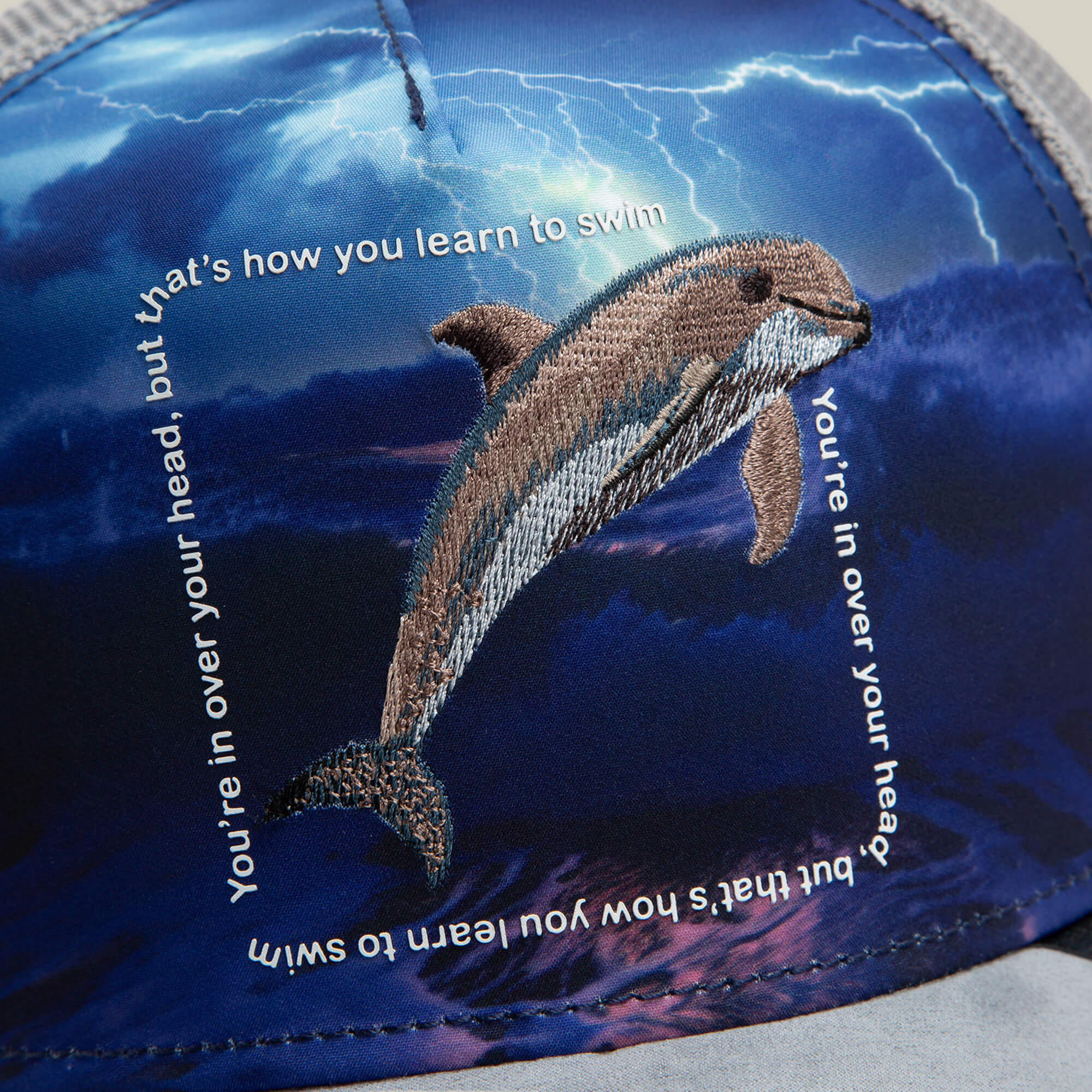 A close-up of the Goorin Bros. "Through the Storm - Dolphin" cap, featuring an embroidered dolphin and motivational text on a blue storm and lightning print—perfect inspirational apparel.