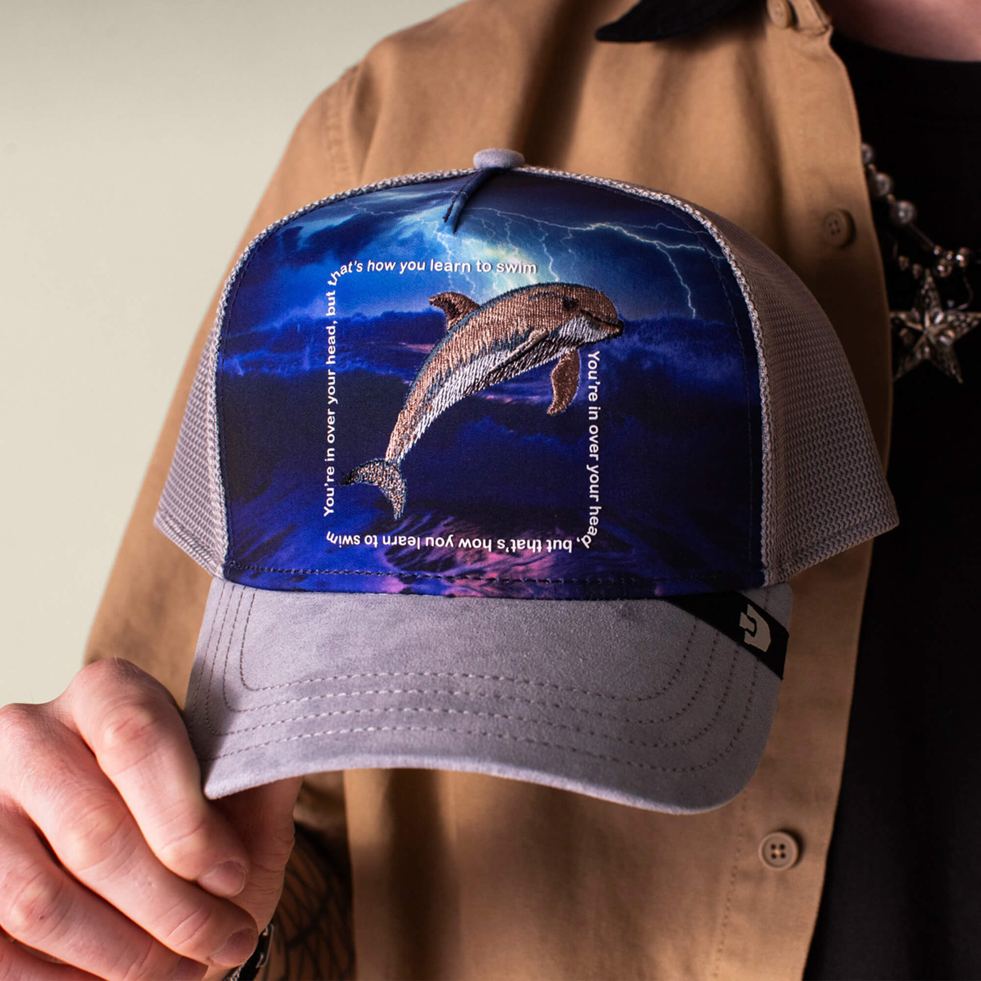 Someone holds the Goorin Bros. "Through the Storm - Dolphin" hat, featuring a gray dolphin graphic and the quote, "You're in over your head, that's how you learn to swim," printed around the image.