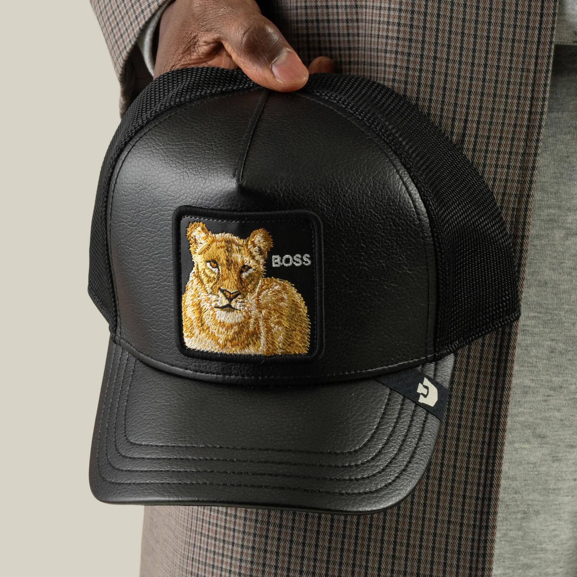 A person holds the Goorin Bros. Faux Leather Lioness trucker hat, featuring black faux-leather, breathable mesh panels, and a lioness patch labeled “BOSS” on the front. The person is wearing a plaid coat and gray shirt.