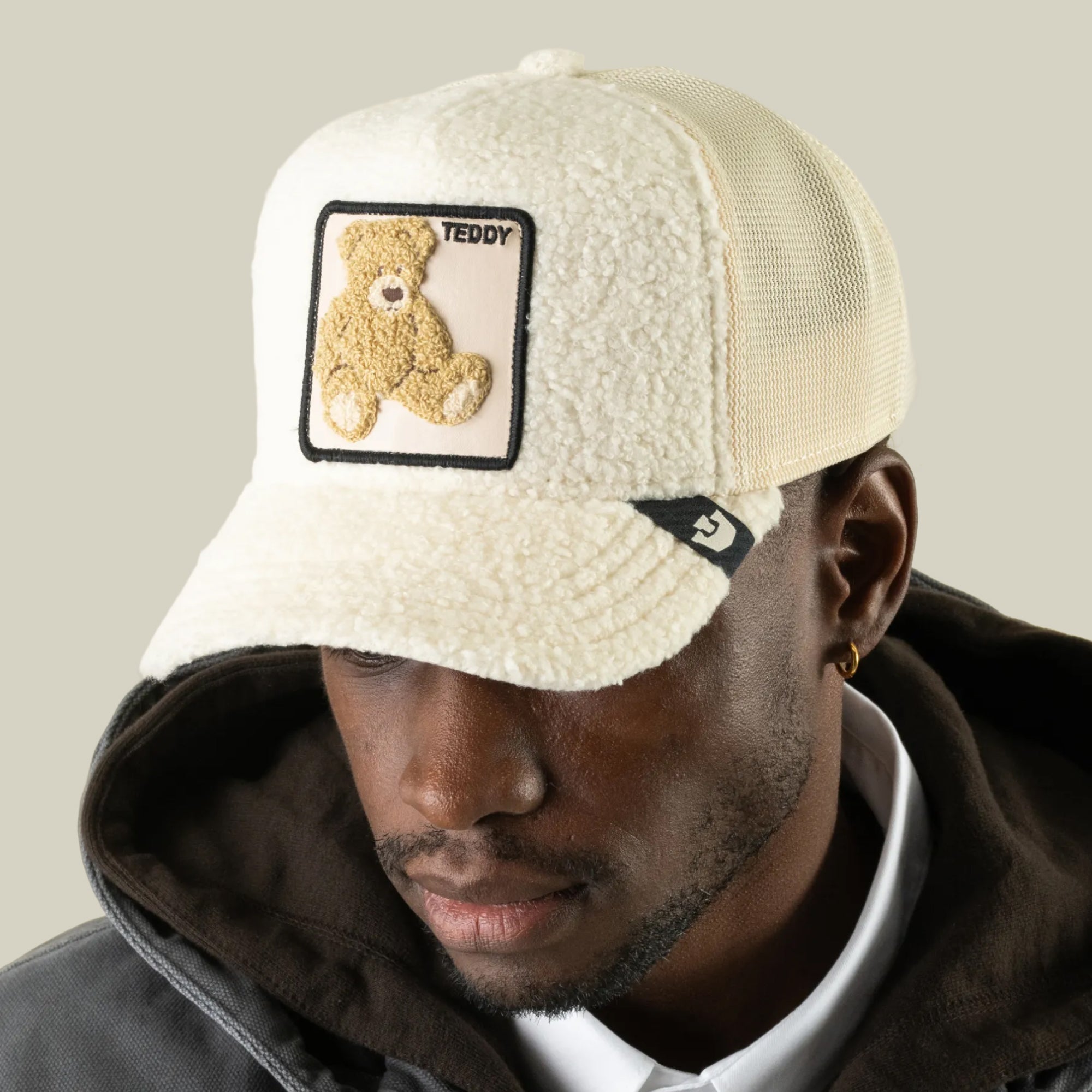 A man wears the Goorin Bros. Double Stuffed - Teddy cream sherpa trucker hat, distinguished by its plush teddy patch and “TEDDY” lettering. Paired with a dark hoodie and white collared shirt, the hat’s breathable mesh back enhances the comfort of his casual, layered style.