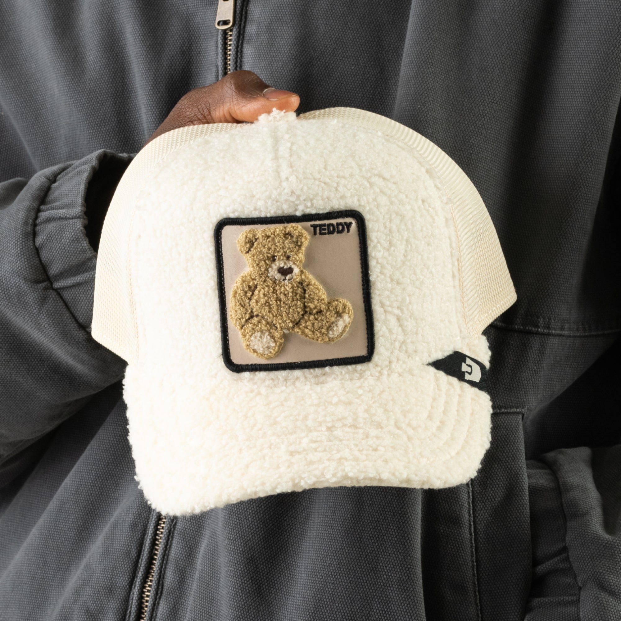 A person in a gray jacket holds the Goorin Bros. Double Stuffed - Teddy sherpa trucker hat featuring a plush teddy patch and the word “TEDDY” on the front.
