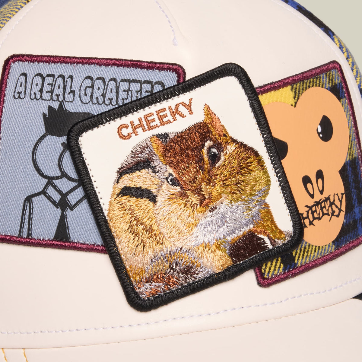 The Goorin Bros. Cheeky trucker hat features a cream and white crown with burgundy-trimmed embroidered patches, including a winking squirrel with "Cheeky" text and humorous character graphics.