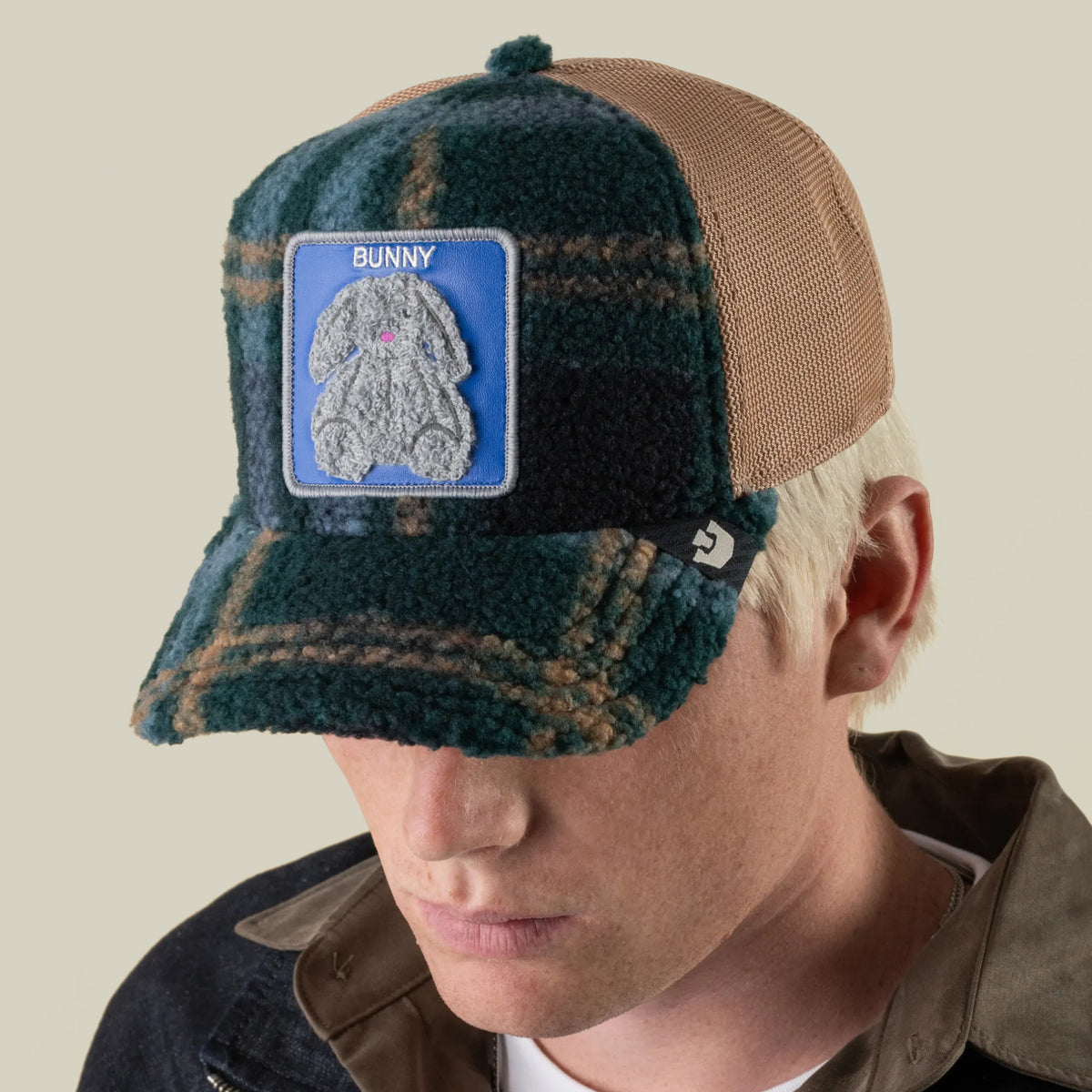 A person wearing the Goorin Bros. Double Stuffed - Bunny trucker hat, featuring a green and brown plaid fleece design, an adjustable snapback, and a blue patch with a plush bunny illustration and the word “BUNNY.”.