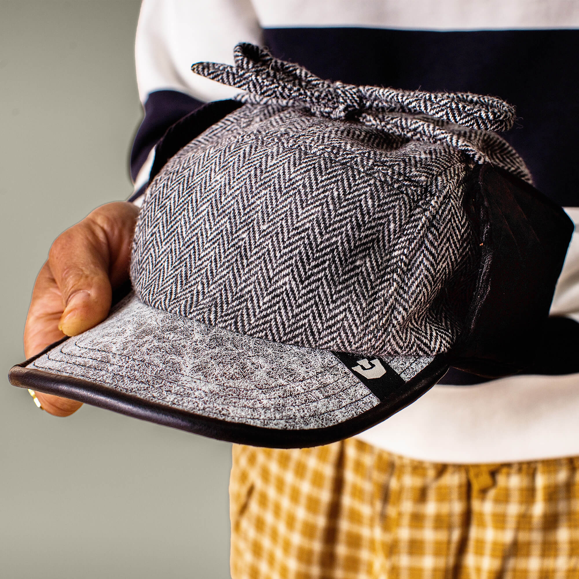 A person holds the Goorin Bros. Frazier Lake herringbone deerstalker hat with a leather brim, paired with a striped top and yellow plaid pants.