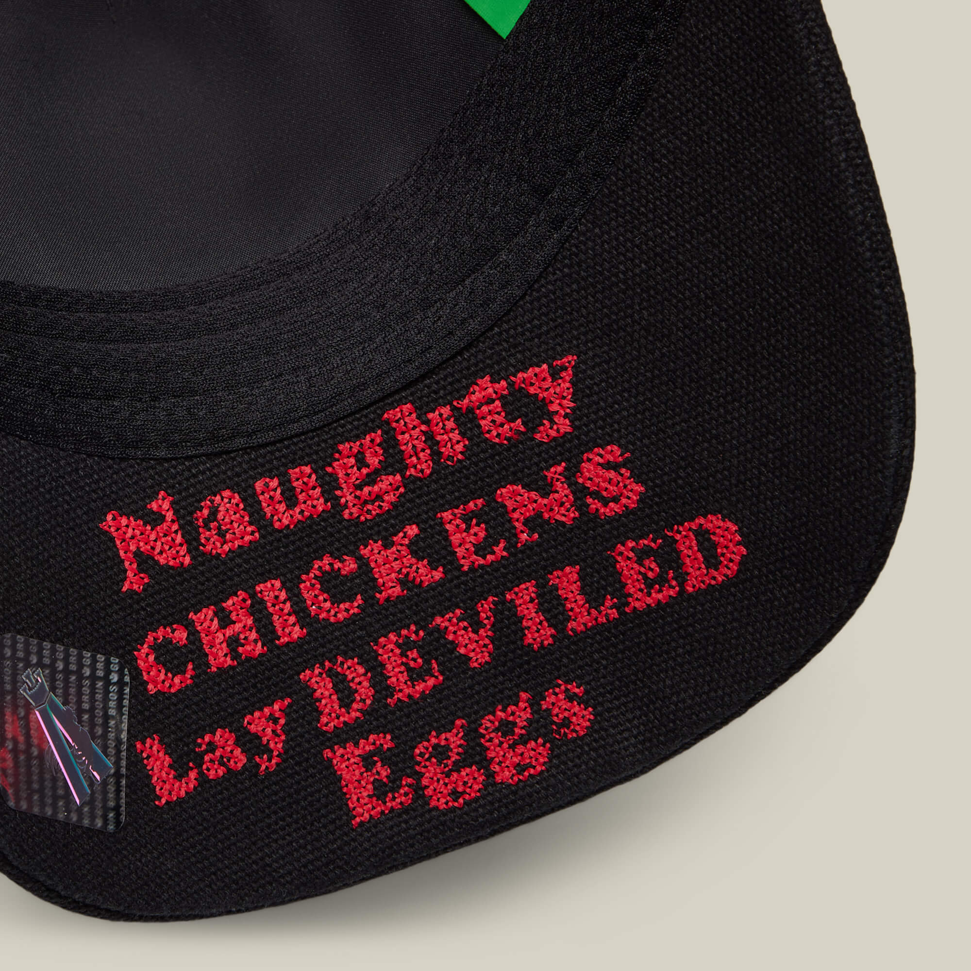 Close-up of black fabric with red embroidered text, "Naughty CHICKENS Lay DEVILED Eggs," from Goorin Bros.’ Middle of No Where collection—ideal for fans of home cooking or fried chicken.