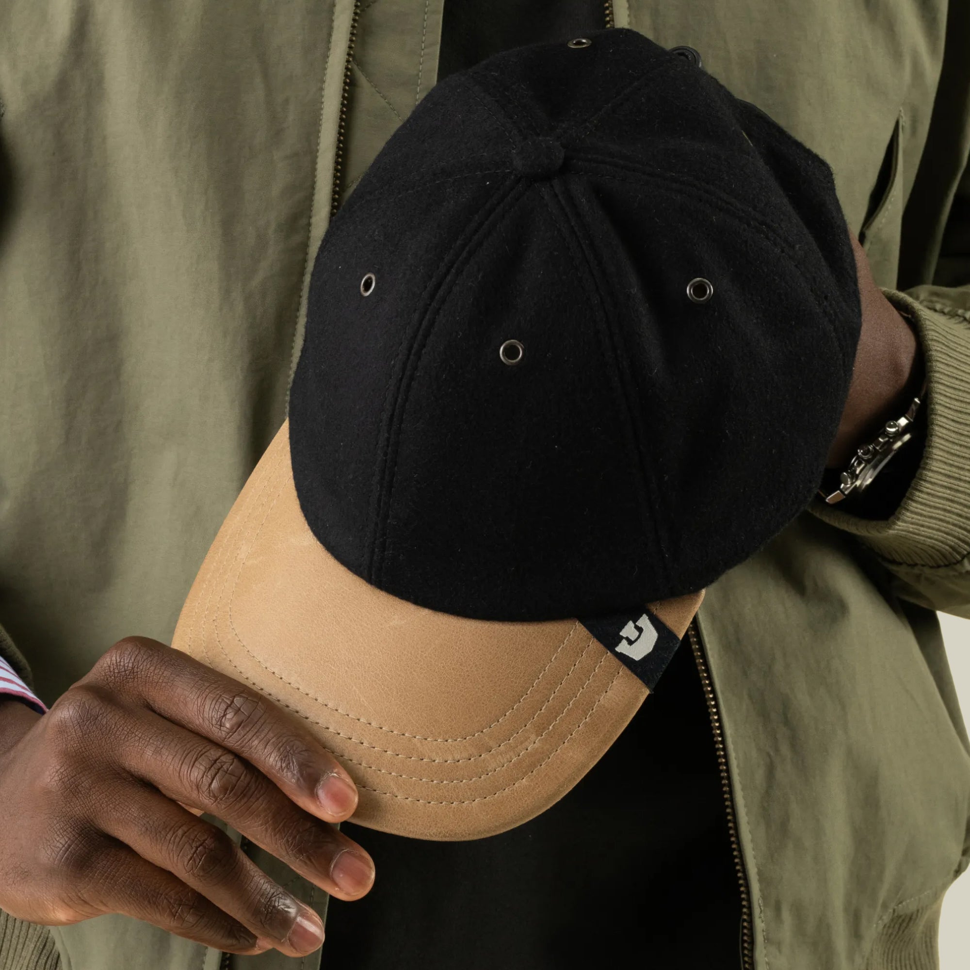 A person wearing an olive green jacket holds a Goorin Bros. Bixby wool leather baseball cap by the brim, with the hand-sewn-in-the-USA logo tag clearly visible.