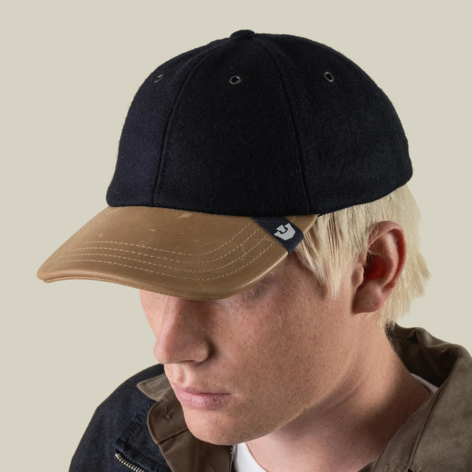 A person wearing the Bixby cap by Goorin Bros.—a hand-sewn black and tan wool leather baseball cap made in the USA—featuring a small logo tag on the brim, looks downward against a plain background.