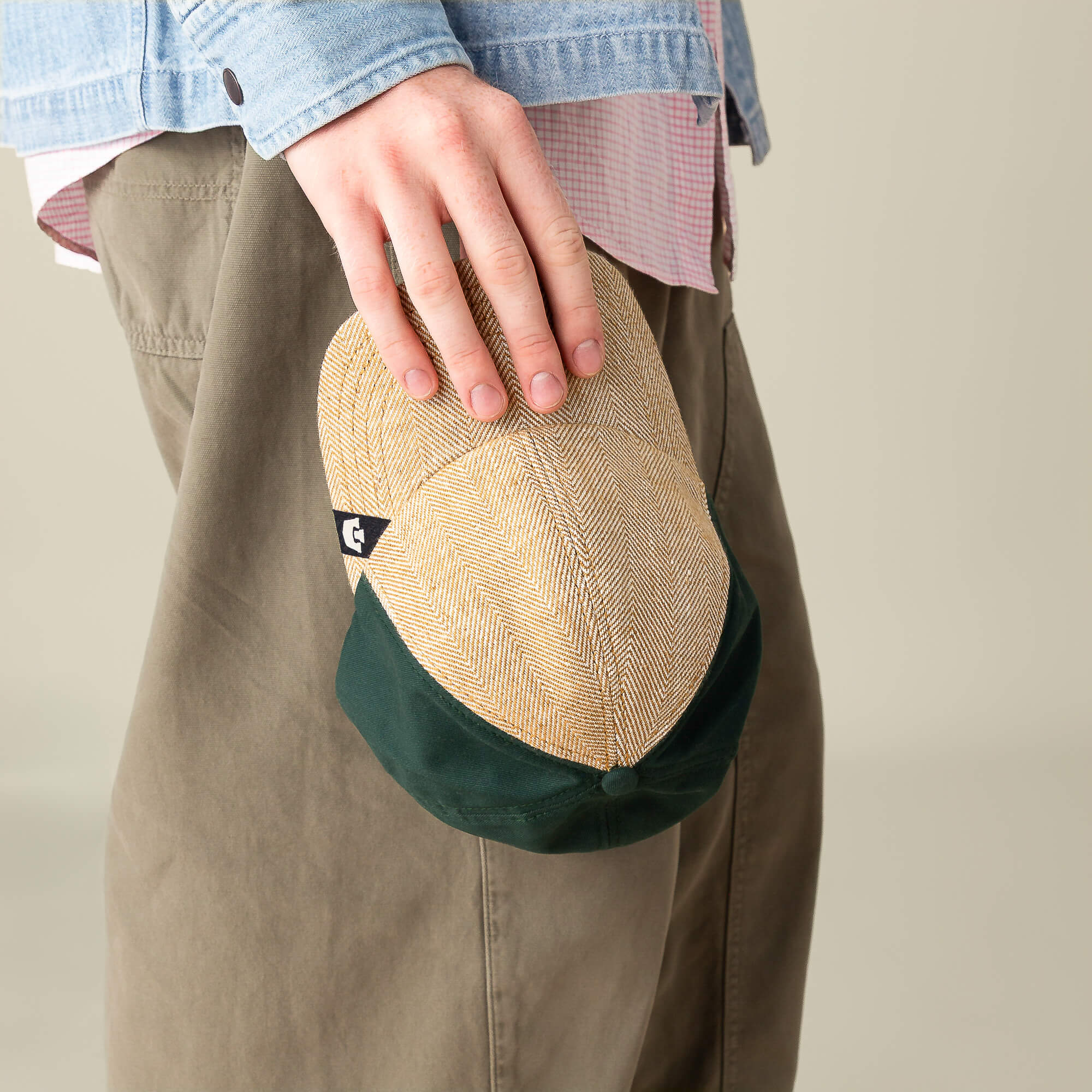 A person holds the Zig Zag Tailored Baseball cap by Goorin Bros. at their side, dressed in olive pants, a pink shirt, and a blue denim jacket.