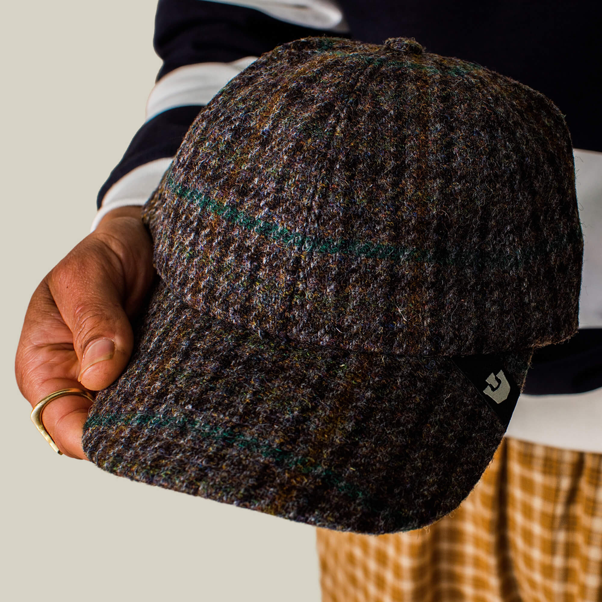 A person holds the Goorin Bros. Cozy Tailored Baseball cap in a muted plaid colorway with a small black tag, wearing a striped sleeve shirt and patterned pants.