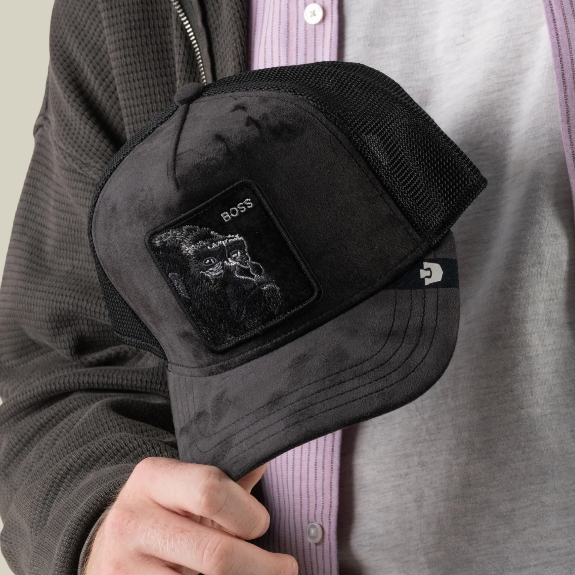 A person holds the Velour Boss trucker hat by Goorin Bros., featuring a black velour finish and a patch with a gorilla and the word “BOSS.” Its breathable mesh design pairs well with their layered gray and lavender shirts.